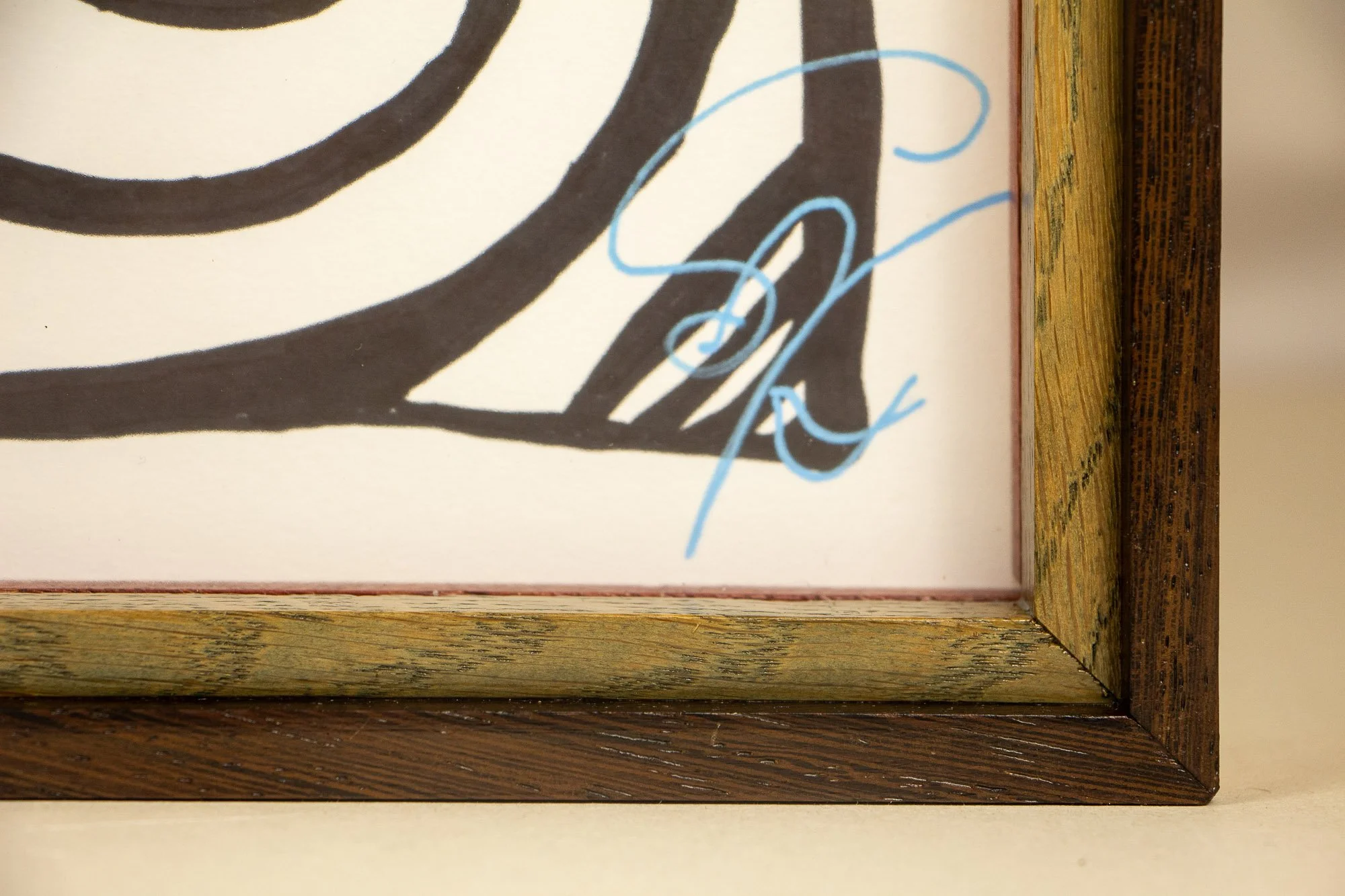 Elusive Cocoon 004 detail and frame: signed lower corner with blue signature on warm white paper, set inside a layered wengé-and-oak frame with olive-toned inner band, orange inner lip, and dark outer rail.