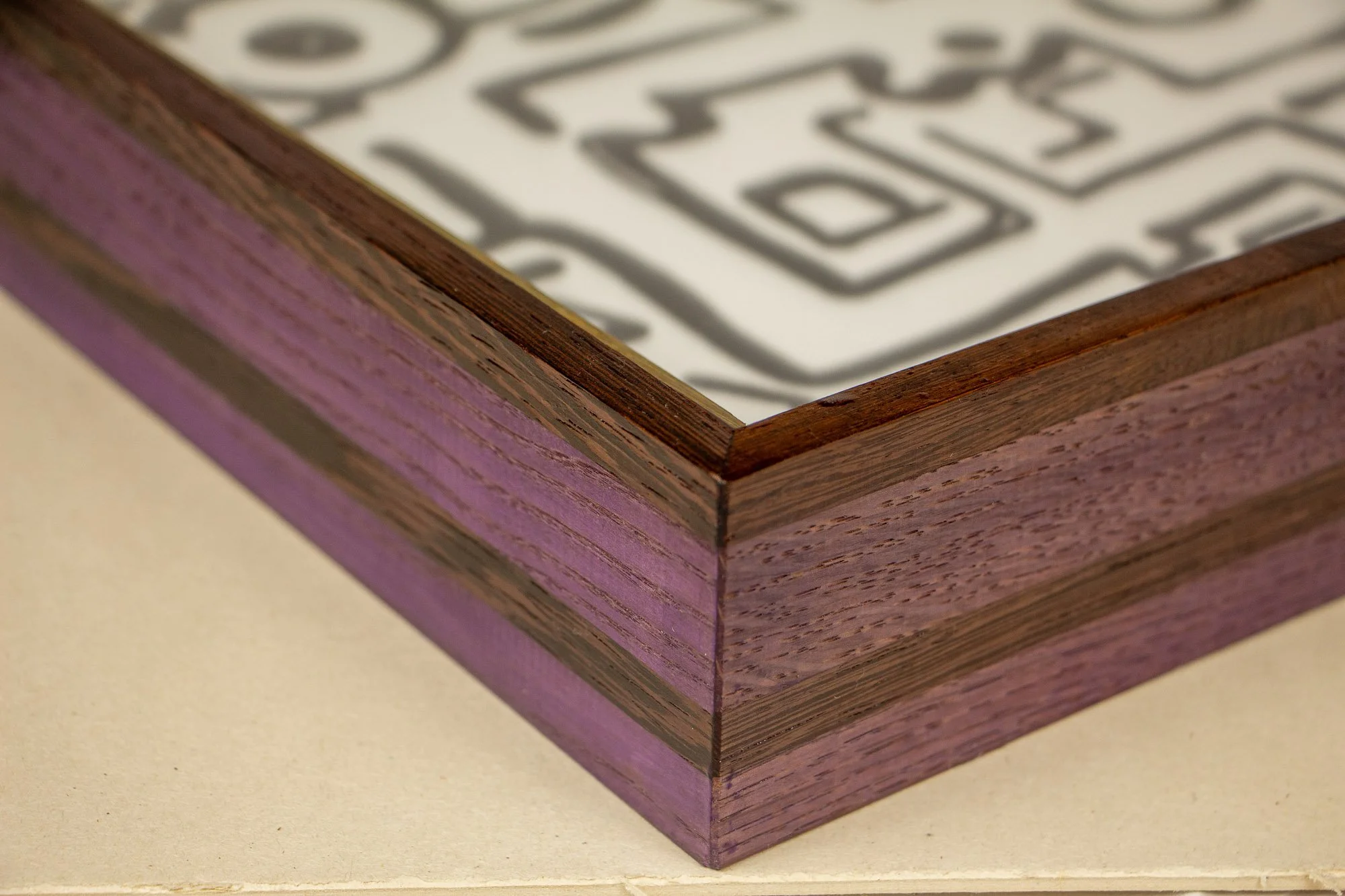 Elusive Cocoon 003 frame: mitered front-corner close-up of the layered wengé-and-oak frame, combining violet-toned oak bands with darker wengé rails around the artwork beneath glass.
