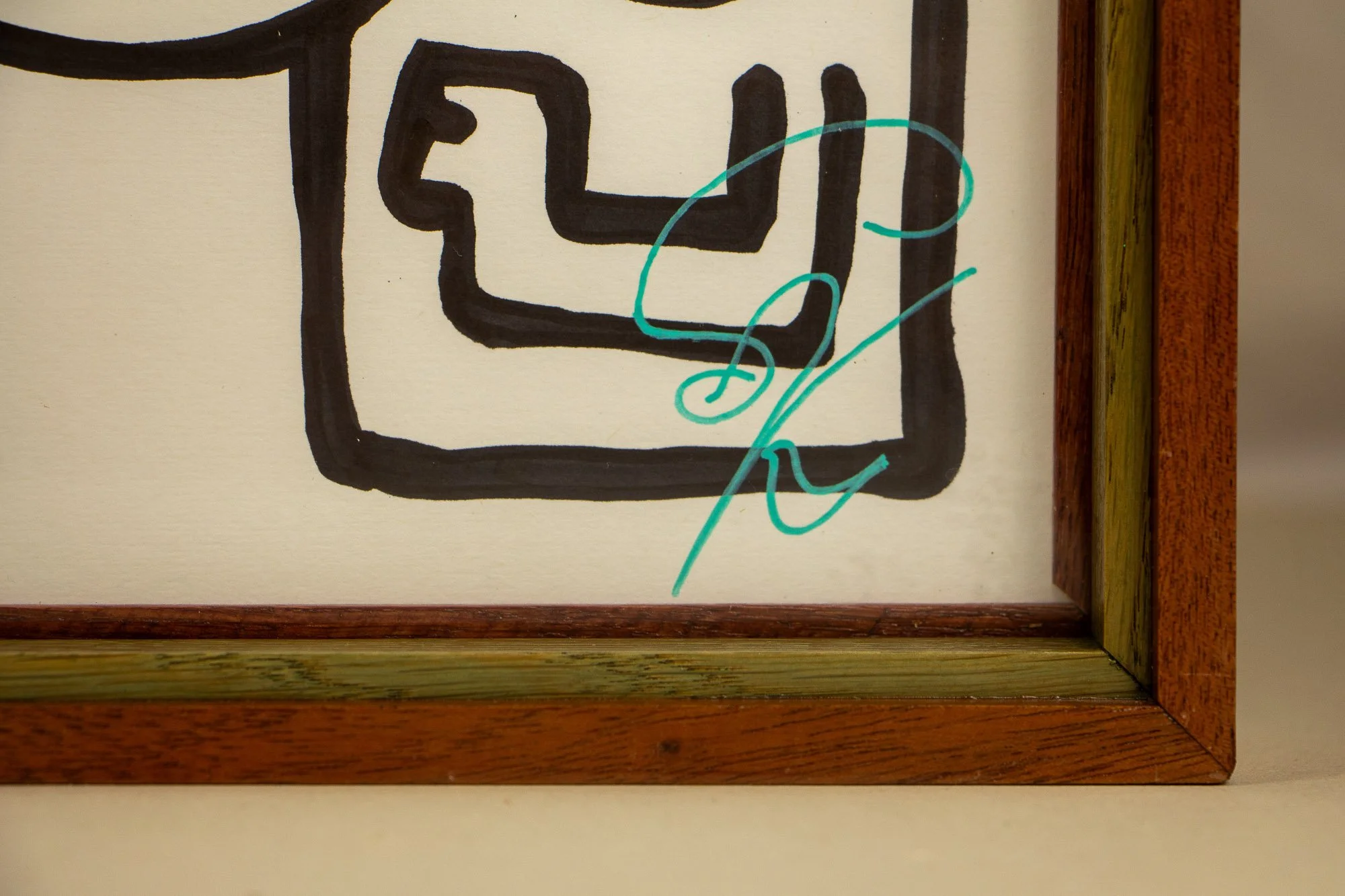 Resonant Syntax 001 frame: lower-right corner detail showing the turquoise signature on paper, bordered by the layered mahogany-and-oak frame in pink, mint, and blue tones