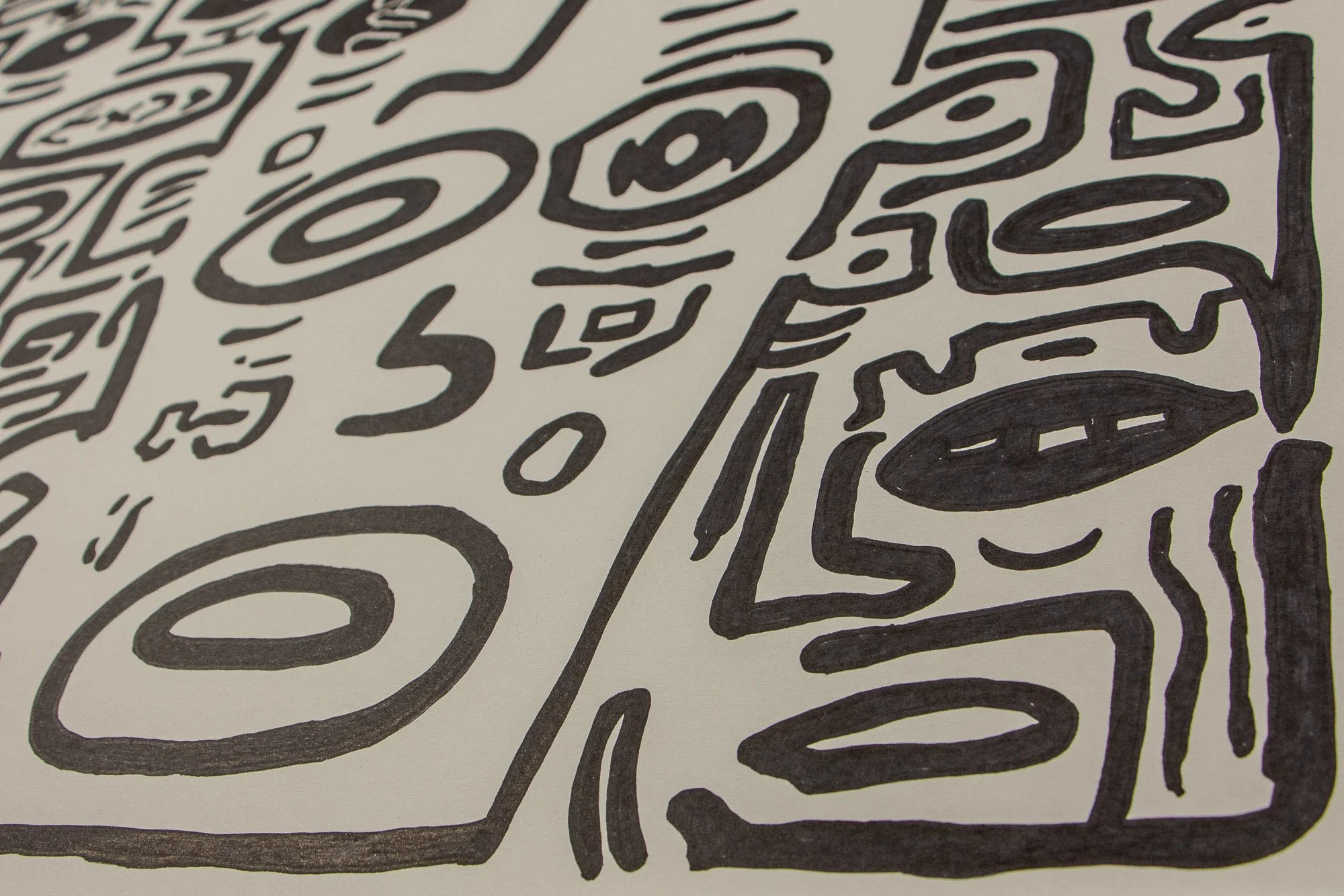 Elusive Cocoon 001 detail: angled close-up of a face-like abstract form with almond-shaped eyes, a rectangular mouth containing arrow-like marks, and surrounding bold black symbols on warm white paper.