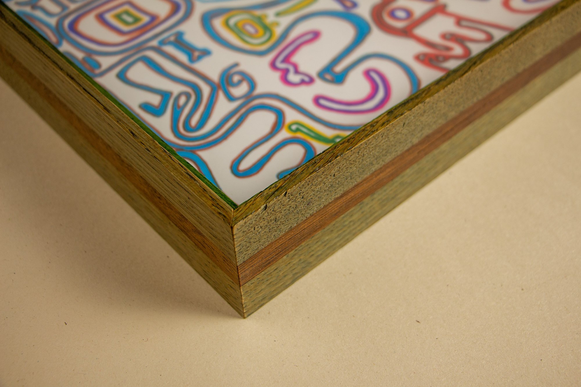 Eclipsing Gravitas 002 frame: angled corner detail of the layered oak-and-mahogany frame, showing olive-green toned oak, a warm mahogany band, a vivid green inner lip, and the multicolored drawing beneath glass.