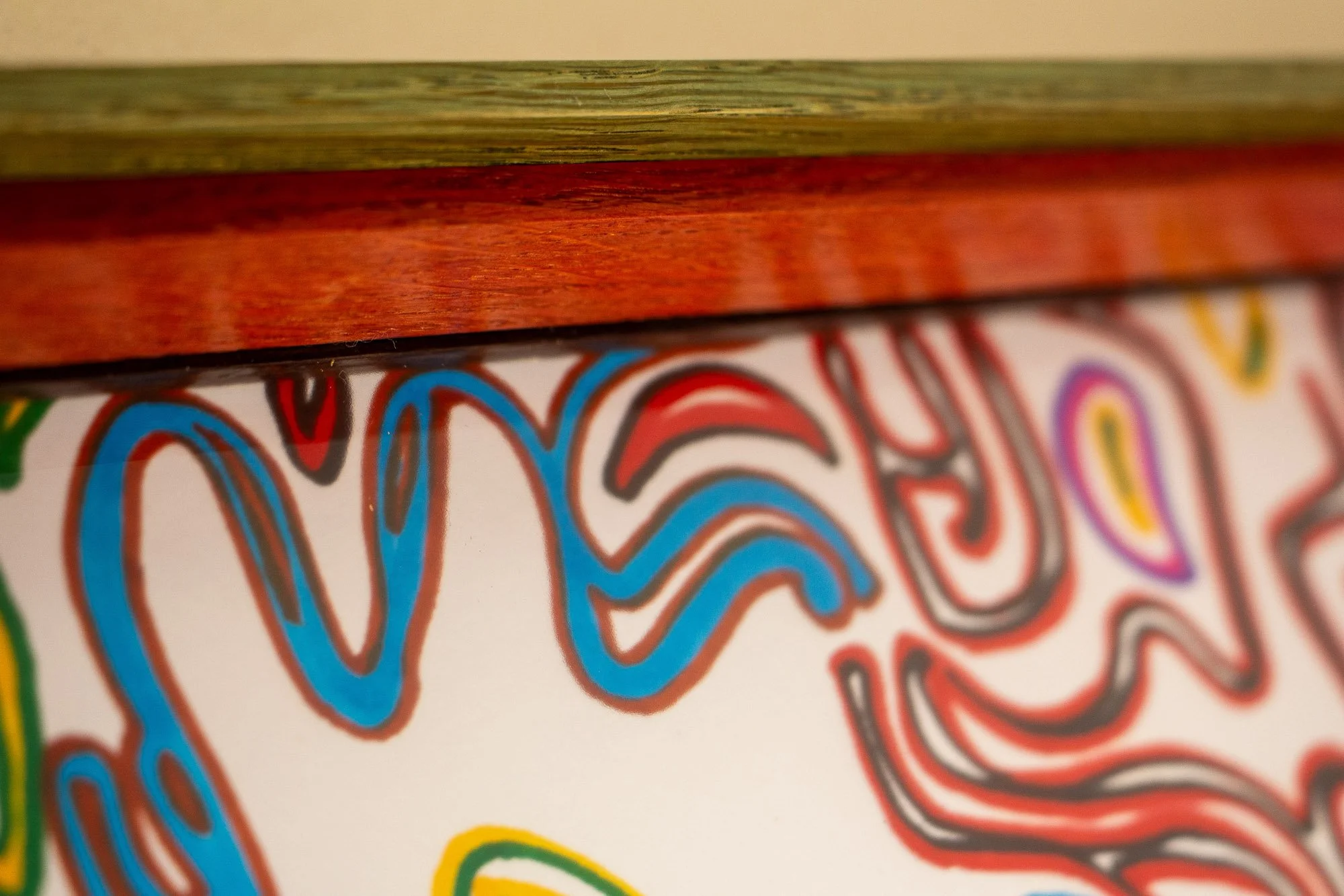 Eclipsing Gravitas 001 frame: edge detail with green-toned oak outer band, warm mahogany inner lip, and the multicolored linework of the drawing softly visible beneath the glass.