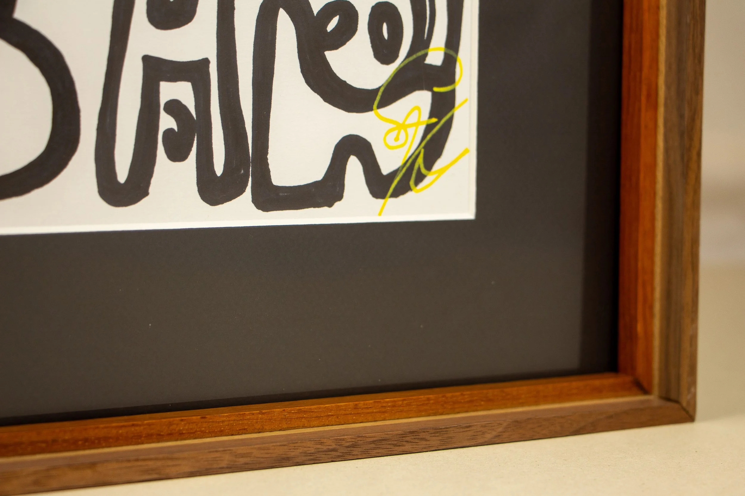 Mêlée of Discovery 002 frame: lower corner detail from the front, showing the black mount, warm inner lip, layered wooden bands, and the lime-yellow signature entering the edge of the composition.