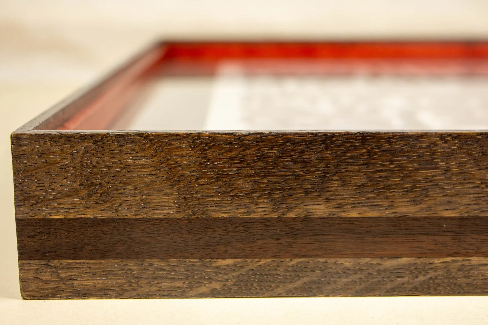 Flux Impromptu 002 frame: long-side profile, dark oak rail with a continuous centered walnut band; glass sits flush at the lip, Granada-red liner in soft focus