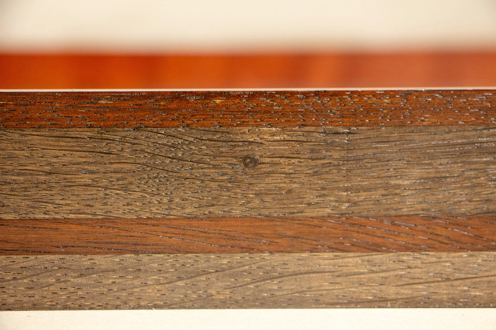 Unseen Signifiers 003 frame detail: straight-on close-up of the side profile, showing a layered dark wood construction with a thin reddish top edge, a muted brown middle field, and a warmer horizontal band running through the center. mahogany, oak an