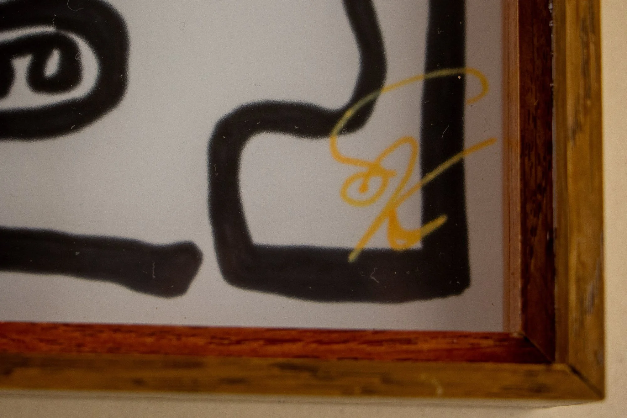 Veiled Alchemist 004 frame detail: lower-right corner close-up of the framed work, showing the reddish inner edge, the warm outer frame, and the artist’s gold signature placed near the margin of the drawing.