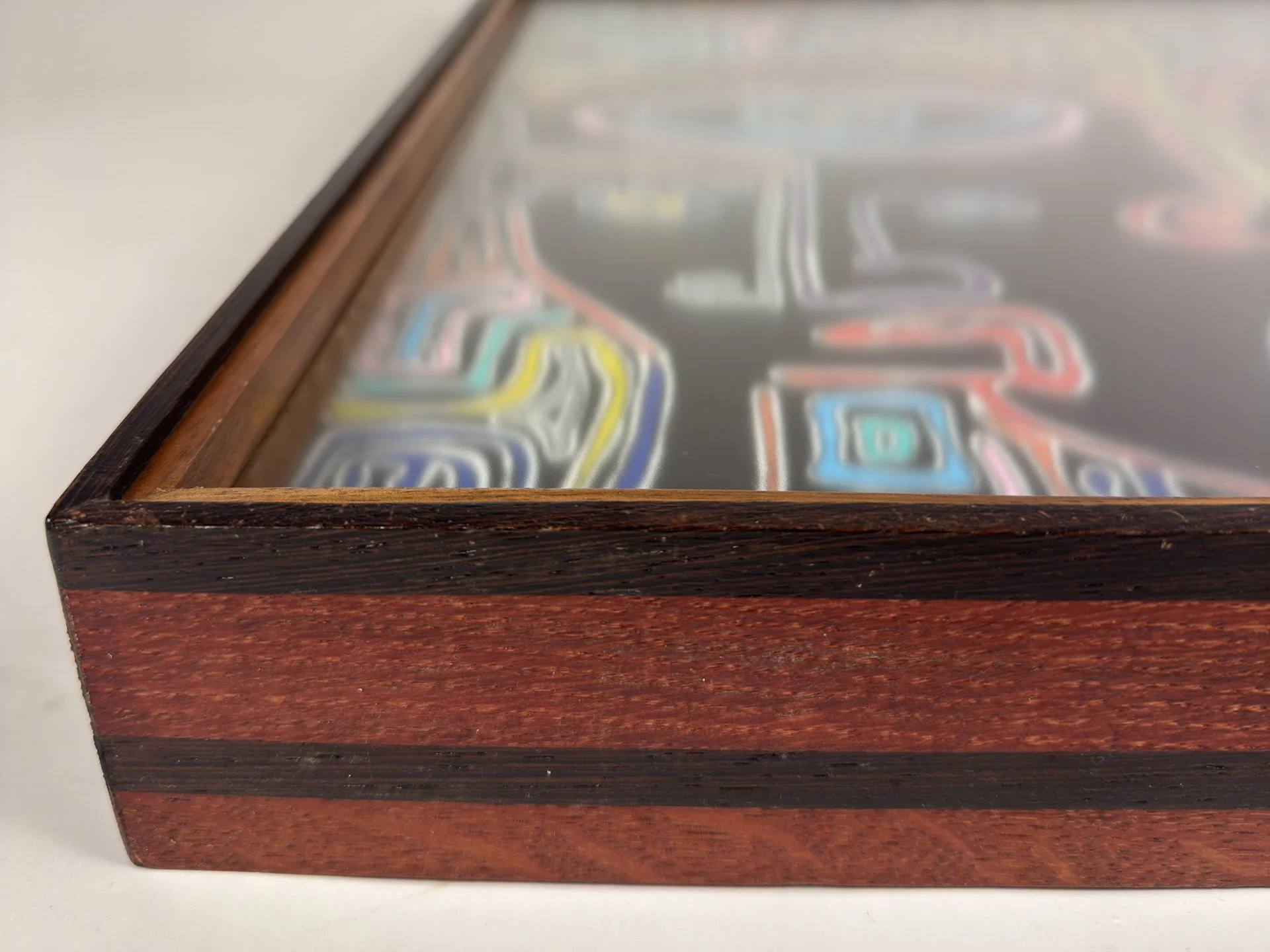 Close-up of the frame’s front corner, highlighting the dark wengé outer edge, warm reddish oak body, and fine walnut inner lip, with blurred sections of the artwork visible beneath the glazing.