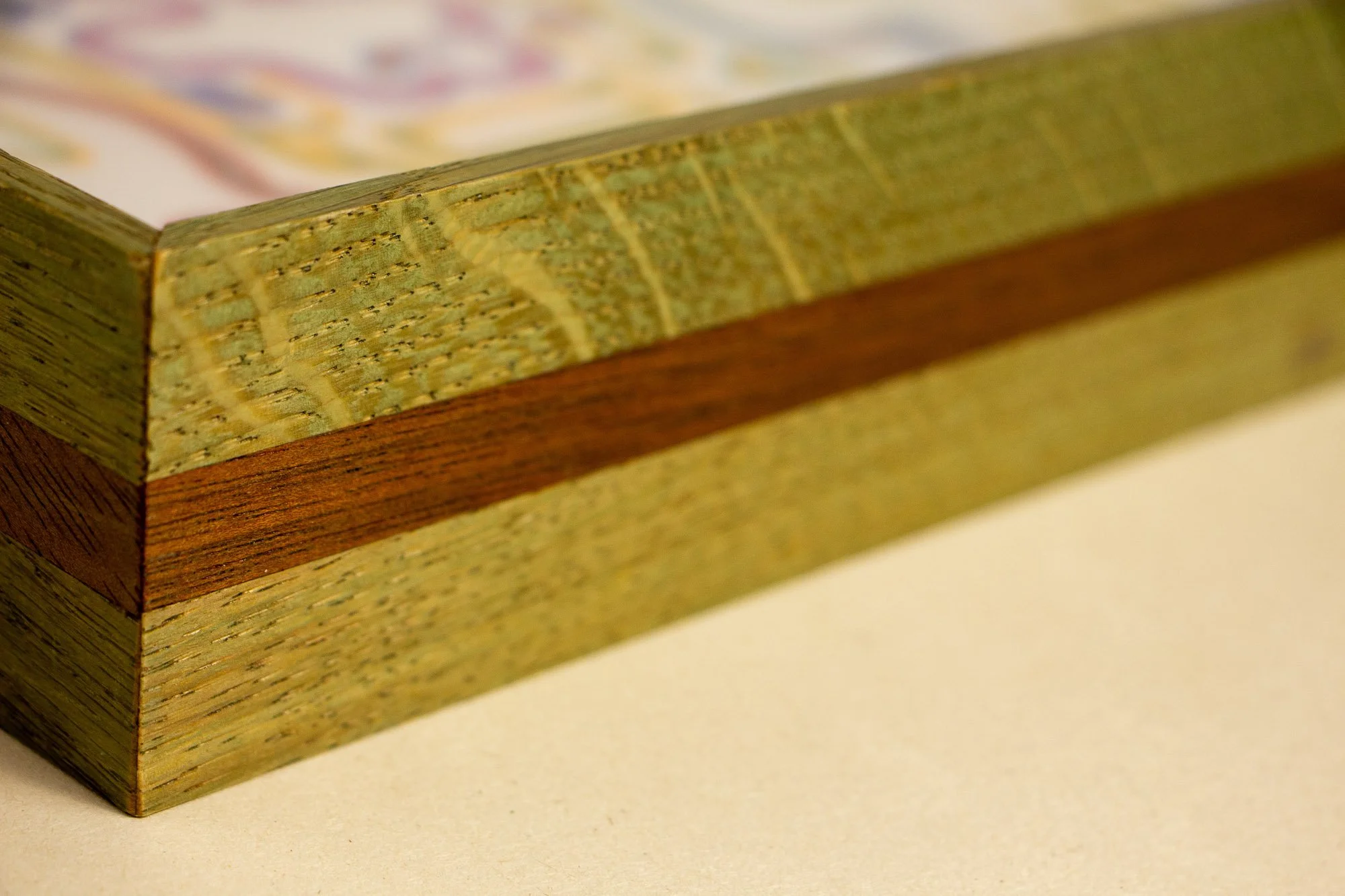 Eclipsing Gravitas 001 frame: angled side close-up of the layered oak-and-mahogany frame, showing green-toned oak faces, a warm mahogany band, visible grain, and a shallow low-profile construction.