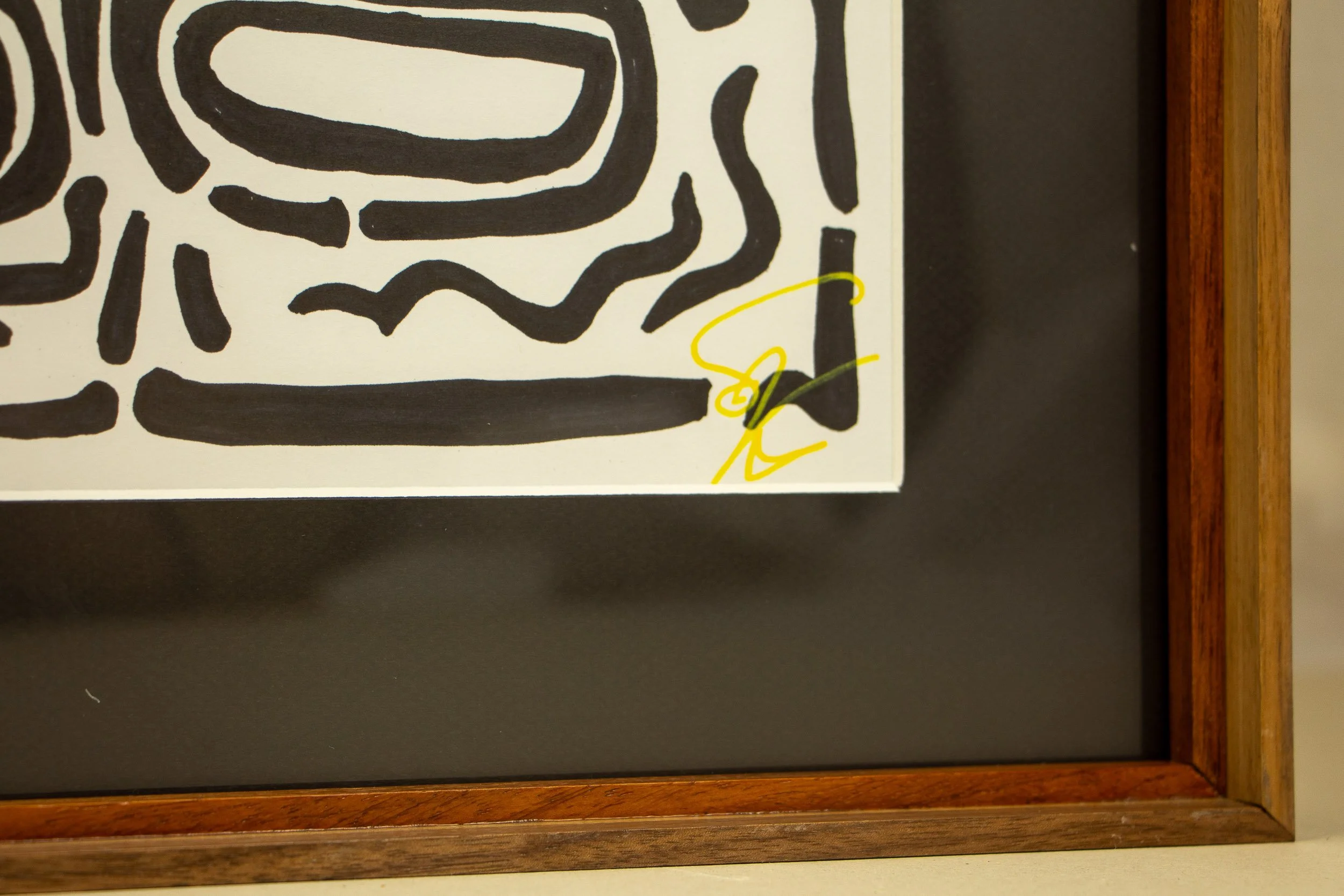 Mêlée of Discovery 004 frame: lower corner detail with the artist’s yellow signature, set against the black mount and bordered by the layered wood frame.