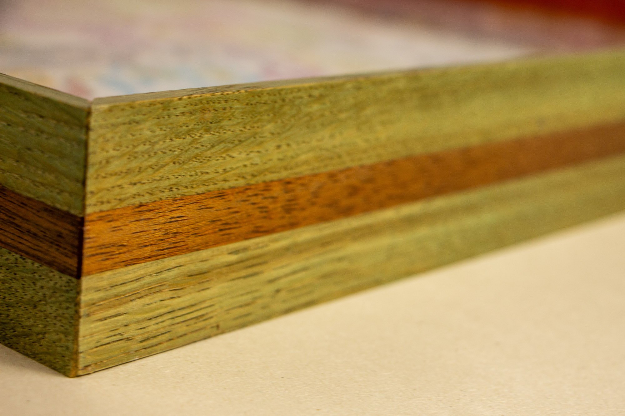 Eclipsing Gravitas 001 frame: angled side detail of the layered oak-and-mahogany frame, showing green-toned oak faces, a warm mahogany band, visible grain, and a shallow profile around the multicolored drawing.