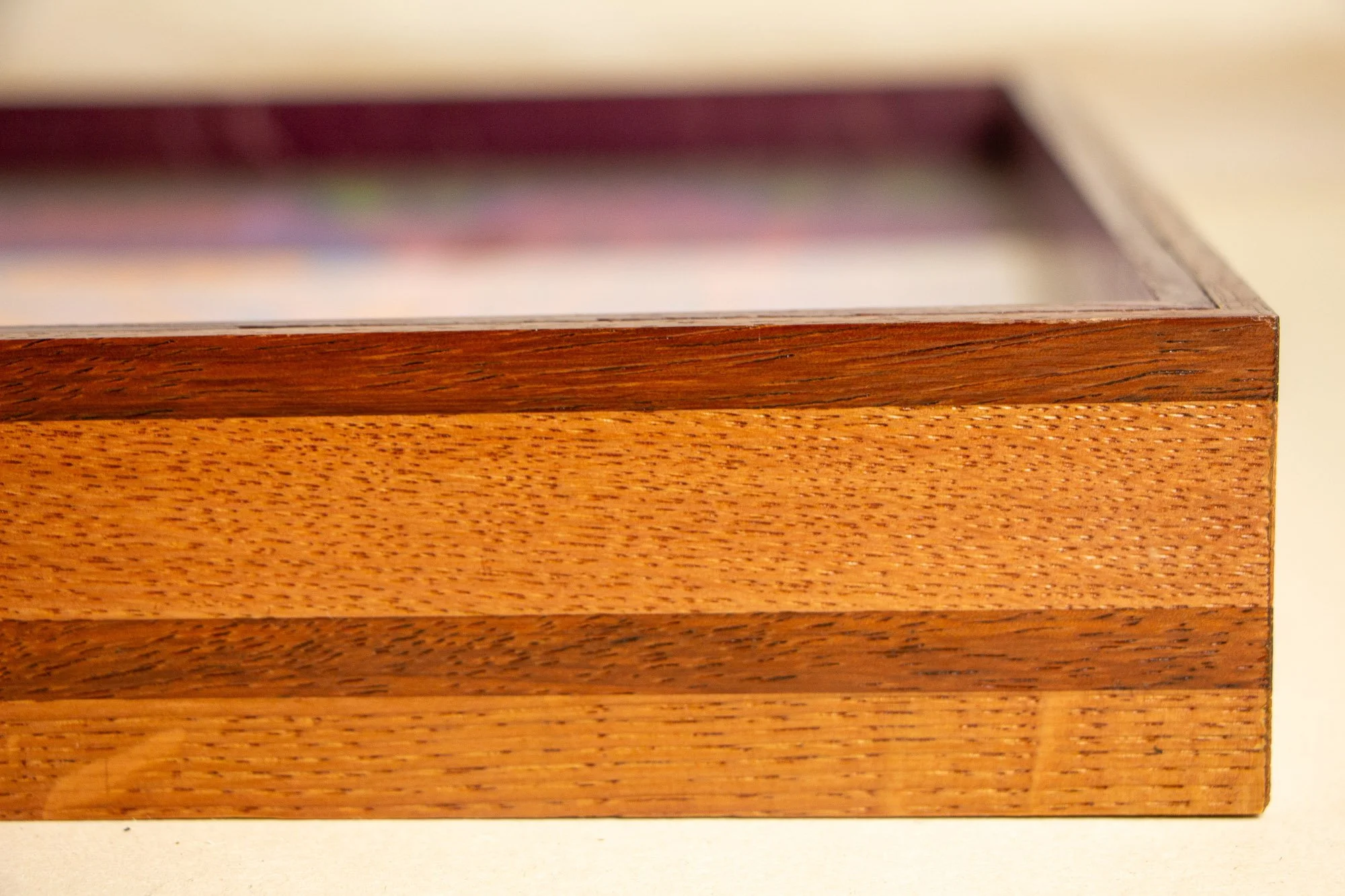 Unseen Signifiers 005 frame detail: front-corner view of the frame, showing the slim upper edge beneath the glazing and the striped wood profile with dark and honey-brown bands meeting at a clean vertical join. mahogany, oak and beech wood.