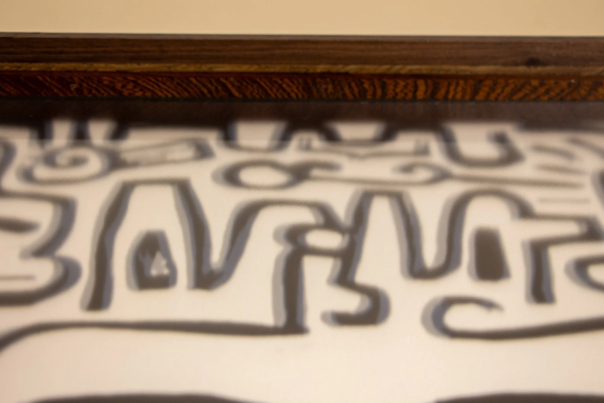 Luminous Parallax 002 frame: close-up along the upper inner edge of the frame, where a richly figured warm-toned inner band sits beneath darker wood, seen above the blurred drawing surface.