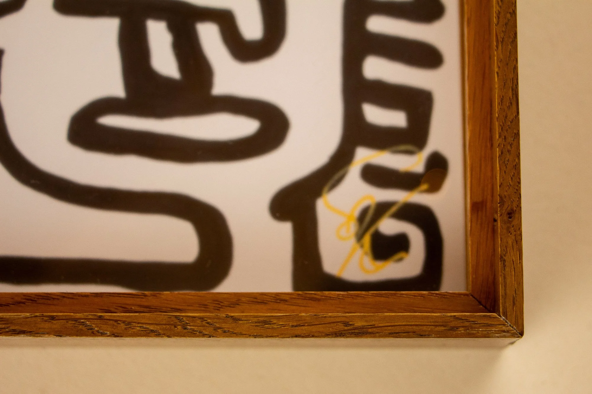 Veiled Alchemist 003 frame detail: lower-right corner close-up of the framed work, showing the mitred joint, the warm wood grain, and the artist’s gold signature near the edge of the drawing.