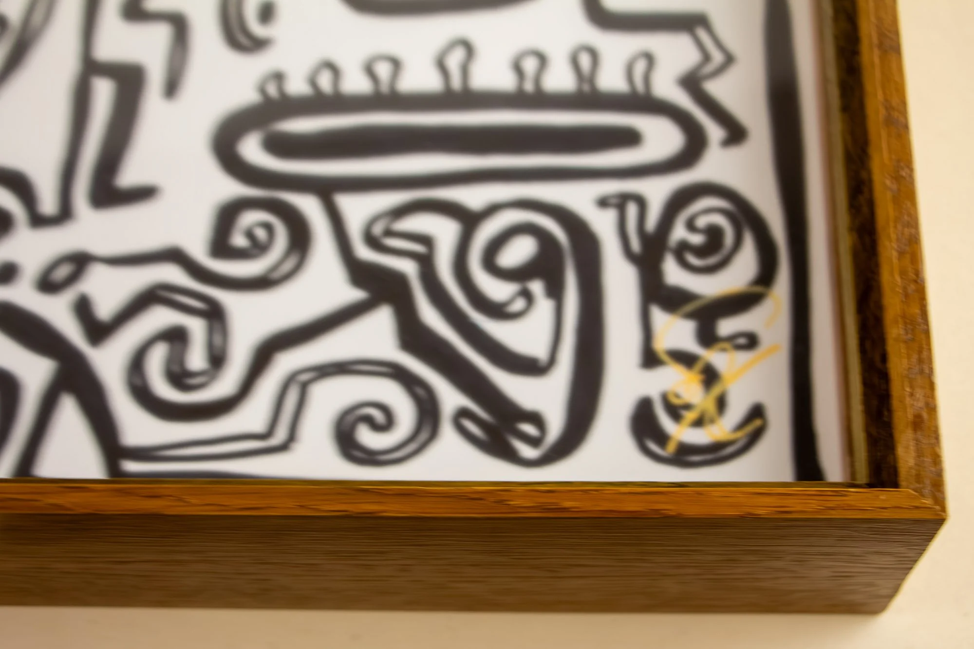 Veiled Alchemist 002 frame detail: lower front view of the framed drawing, showing the gold signature near the bottom right, the warm wood frame, and the artwork held behind glass within a deep, quiet structure.