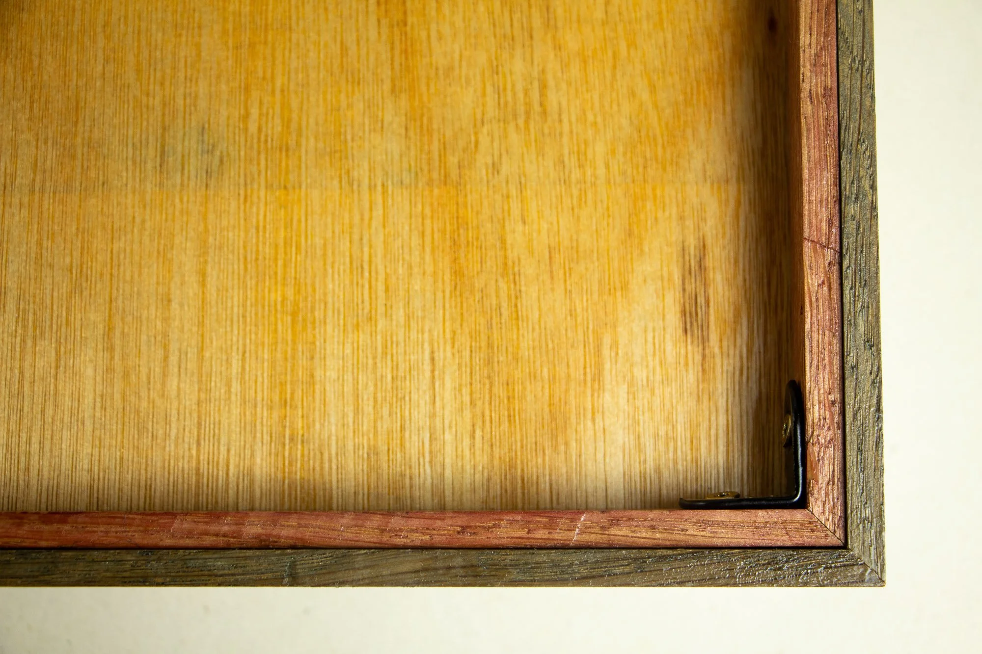 Unseen Signifiers 001 frame detail: reverse corner view of the frame, showing the pale wood back panel, the reddish inner structure, the olive outer edge, and the black corner bracket fixing the construction in place.