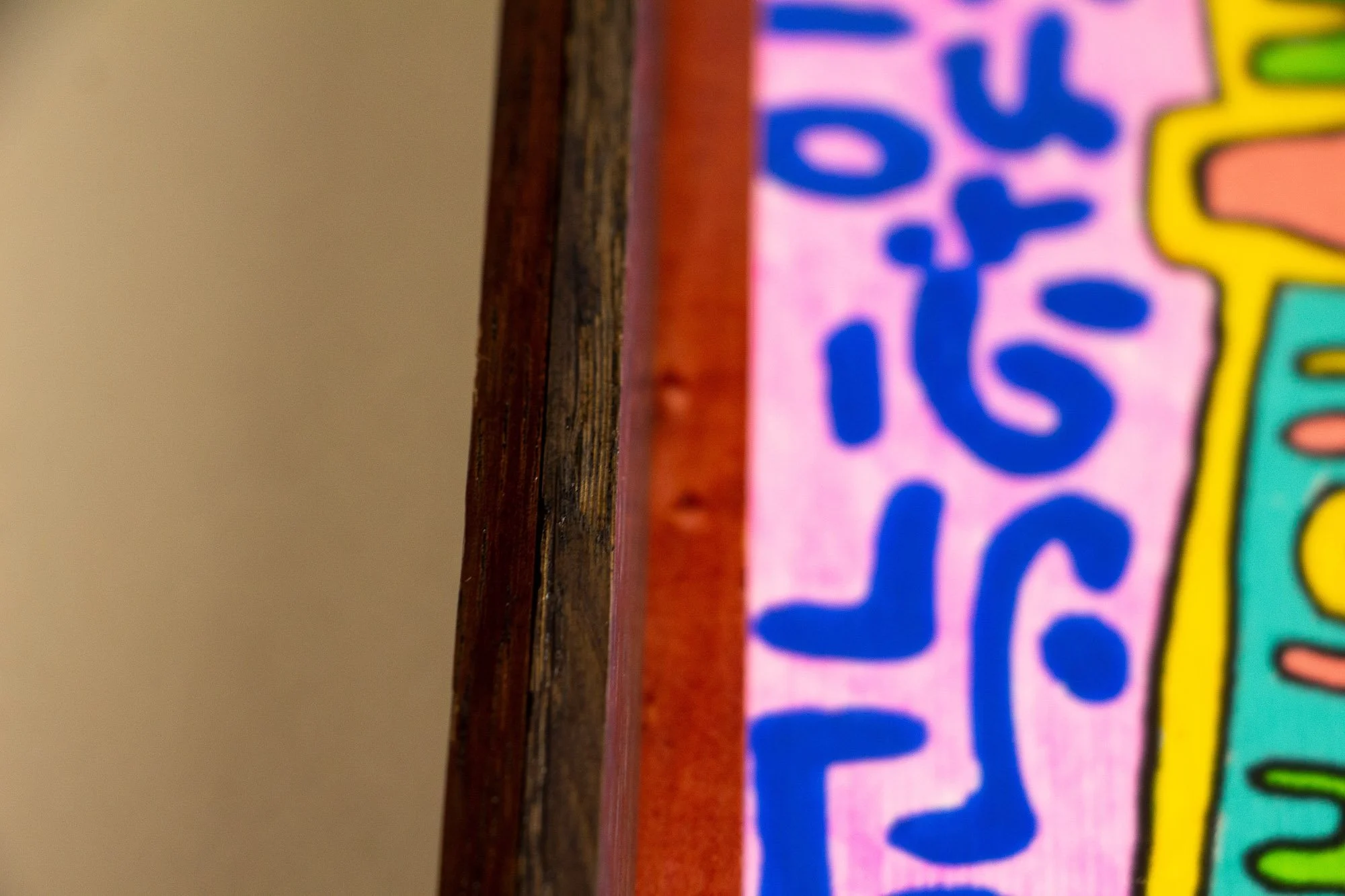  mahogany, oak and beech wood.Unseen Signifiers 007 frame detail: close-up of the frame’s side edge, revealing the dark outer wood, warm reddish inner strip, and the pink artwork surface with blue symbols behind the glass.