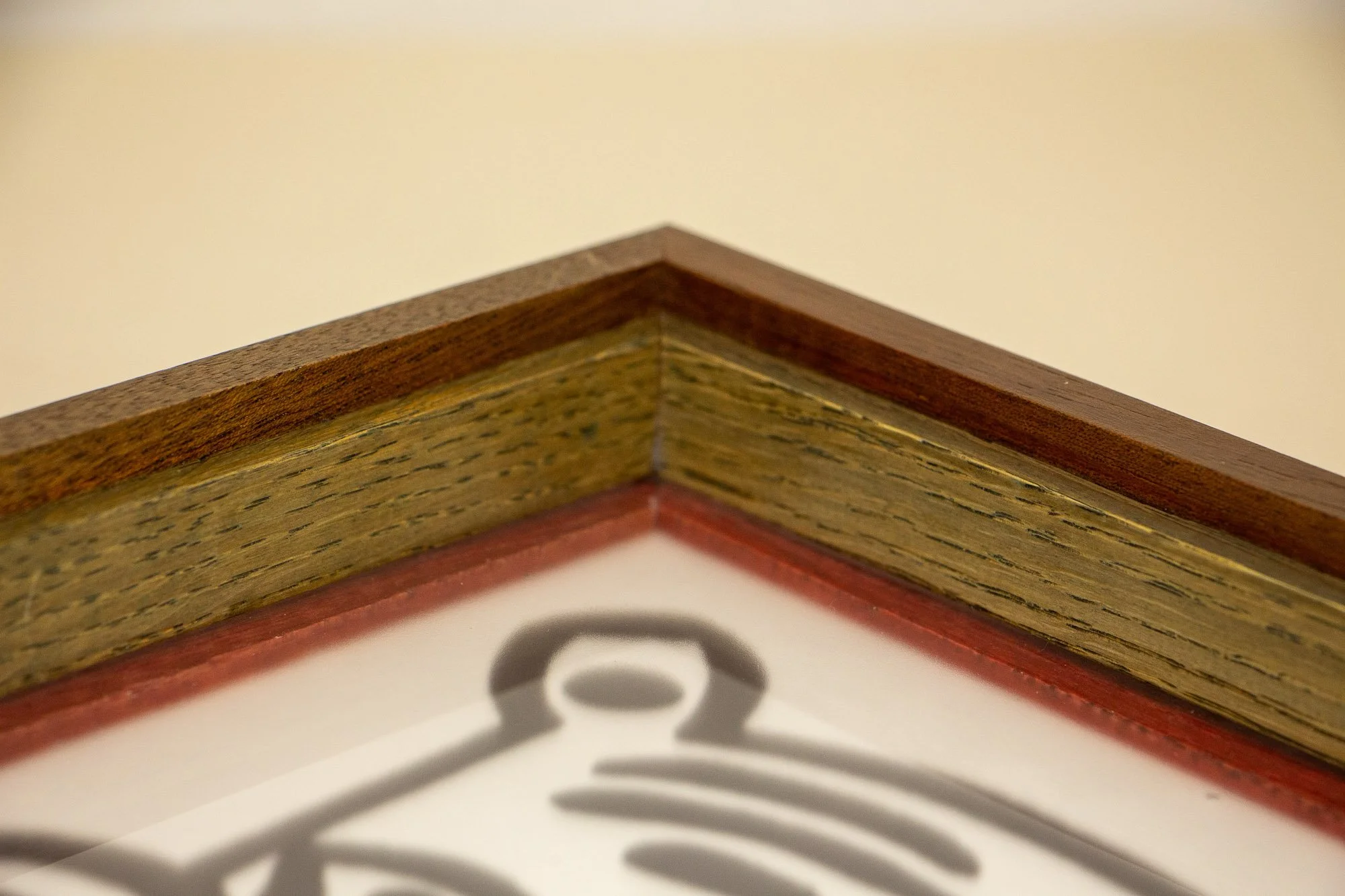 Resonant Syntax 002 frame: upper corner detail with mahogany outer frame, colored oak inner band, and a narrow inner lip bordering the artwork beneath the glass