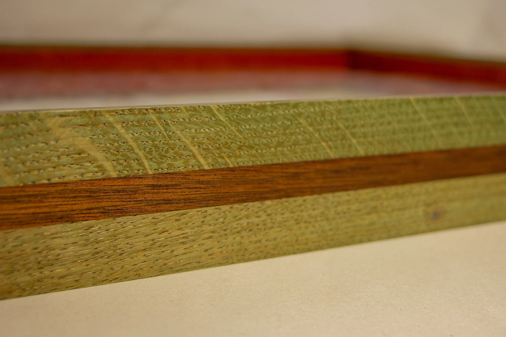 Eclipsing Gravitas 001 frame: frontal close-up of the frame profile with green-toned oak, a warm mahogany band, and a broad low-profile face surrounding the artwork beneath glass.