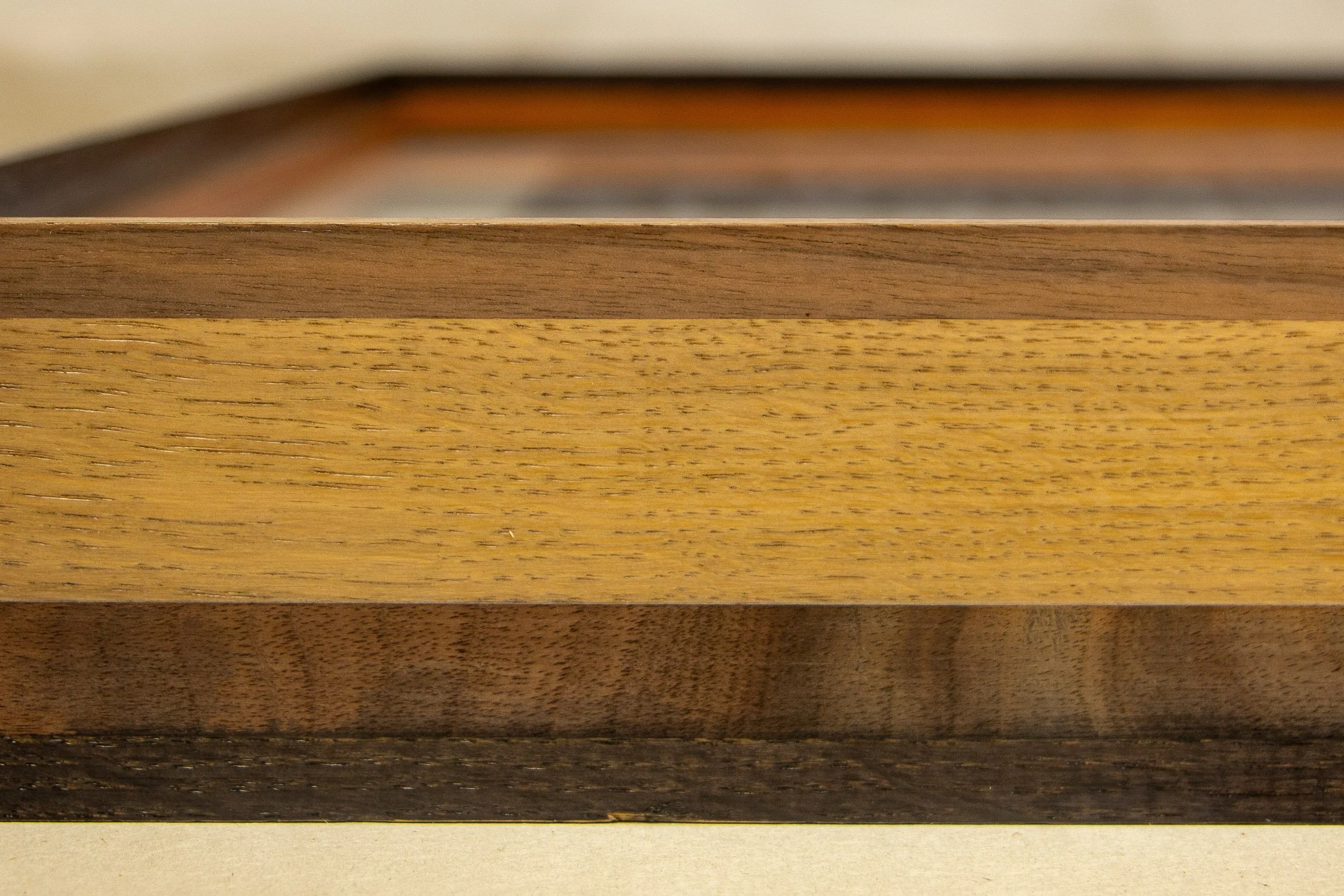 Mêlée of Discovery 003 frame: front corner detail showing the frame’s layered construction, where a light central band is held between darker upper and lower wooden strips.