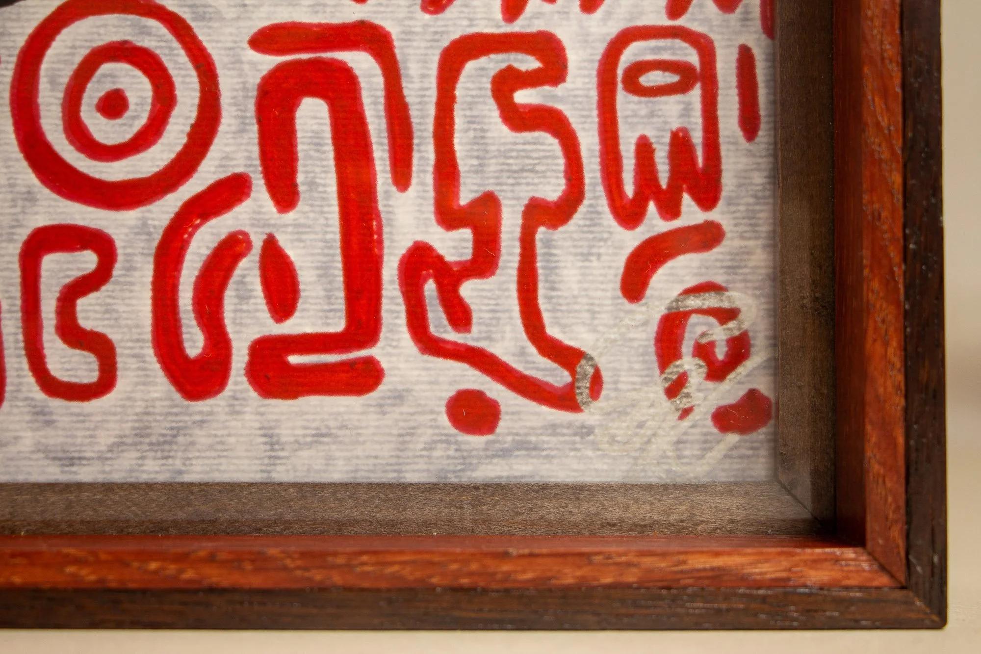 Unseen Signifiers 003 frame detail: lower-right corner of the artwork and frame, with red symbols on the pale ground, the silver signature near the edge, and the red inner lip framed by darker wood. mahogany, oak and beech wood.