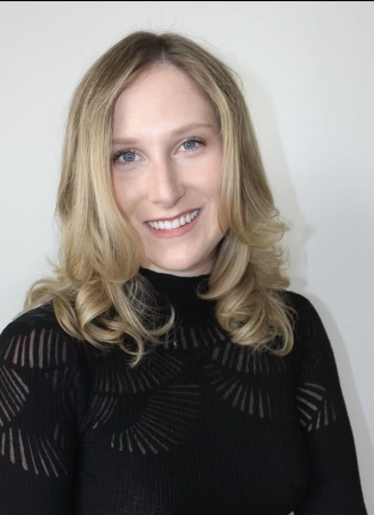 A woman with blonde hair, blue eyes, wearing a black top with a sheer, patterned design, smiling at the camera against a plain white background.