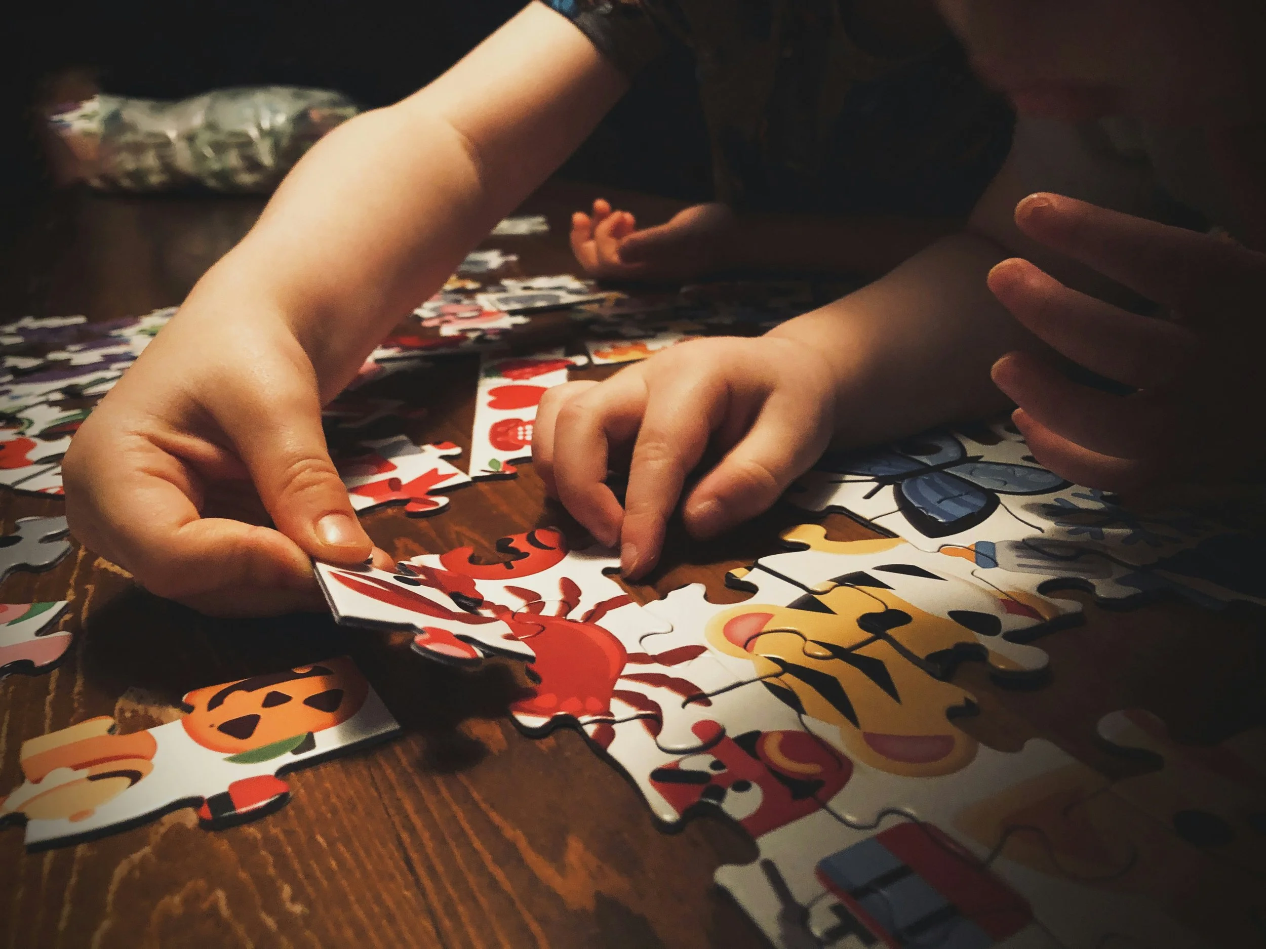 Two children are working together to assemble a colorful jigsaw puzzle on a wooden table. Their hands are visible, sorting and fitting the puzzle pieces, which feature various cartoon-like images.