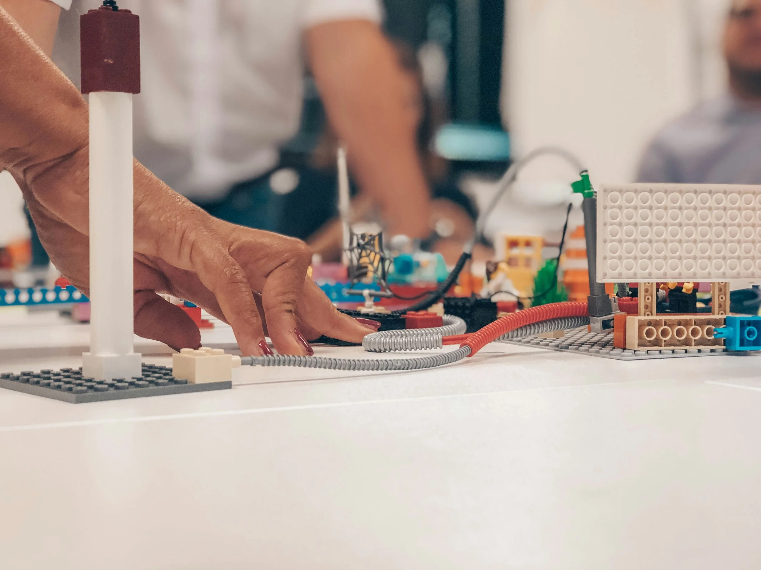Close-up of a hand pointing to a LEGO creation on a table, with blurred people in the background.