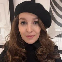 Young woman with wavy brown hair wearing a black beret and black top, smiling at the camera in an indoor setting.