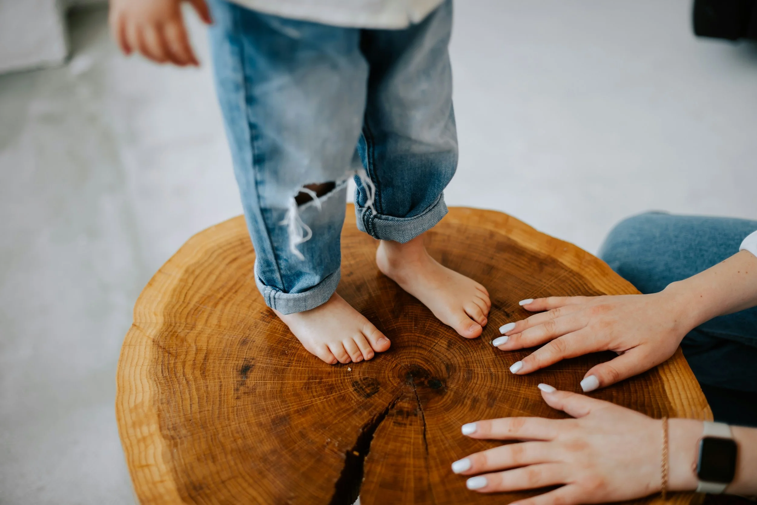 Close-up of a child's feet standing on a wooden table with an adult's hands resting nearby, showing the child's ripped jeans and bare feet.