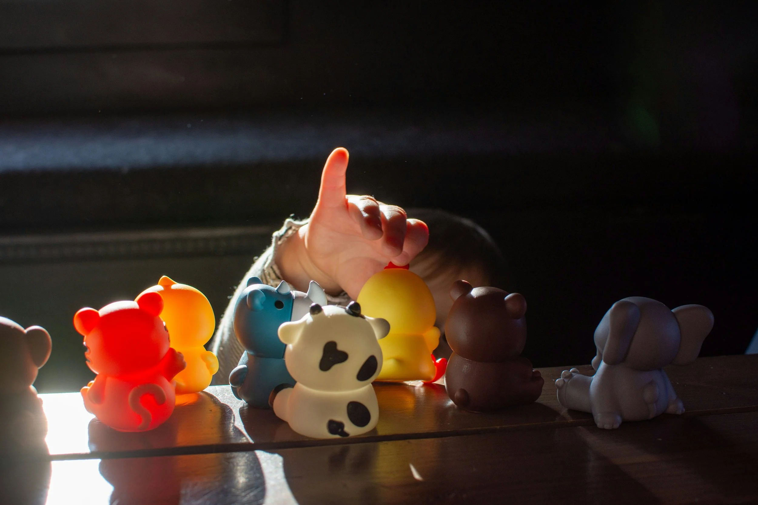 Child's hand reaching over a row of colorful animal toys on a wooden surface, backlit with sunlight creating silhouettes.