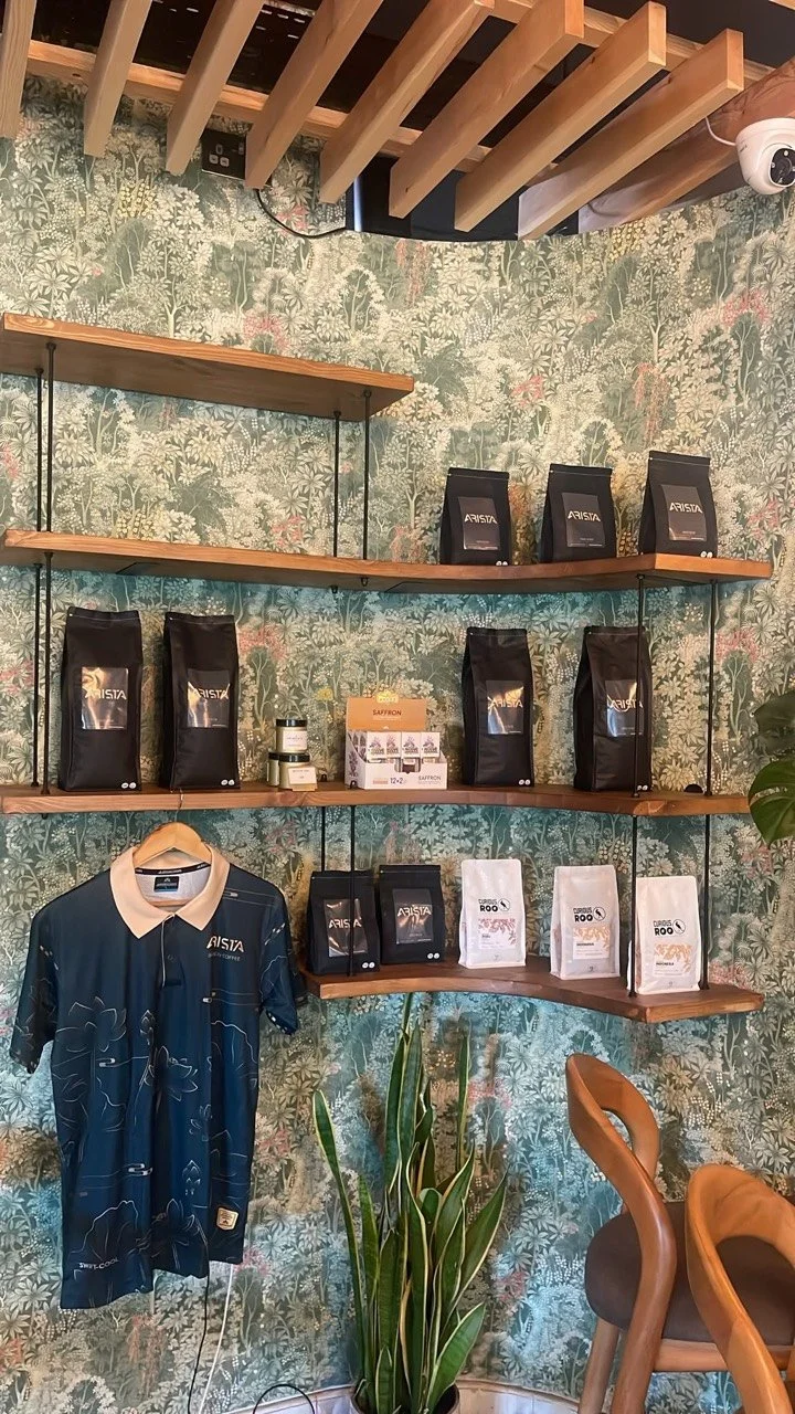 Completed Interior Detail — Timber display shelving and coffee packaging against botanical wallpaper beneath the slatted ceiling.