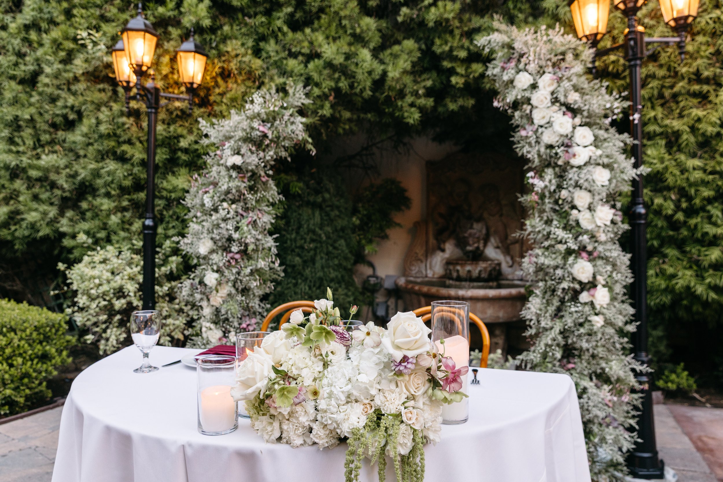 Round table with white tablecloth, floral centerpiece, candles, and glasses outdoors in a garden with greenery and street lamps, decorated for a special occasion or event.