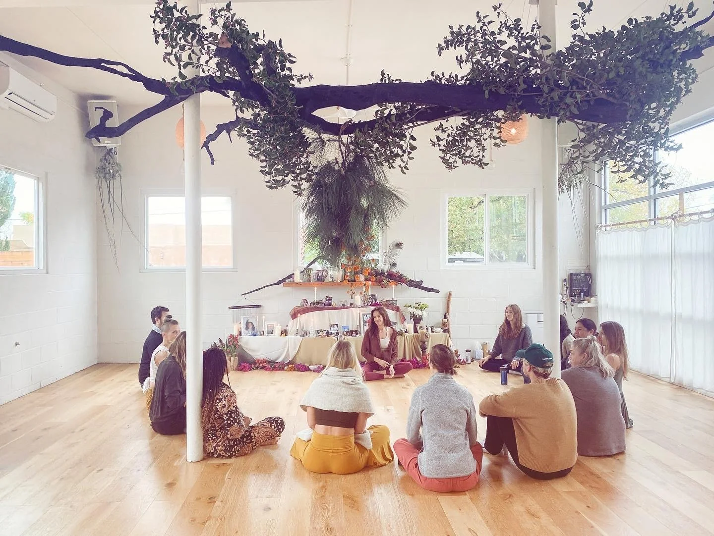 A group of people sitting on the wooden floor in a circle in a bright, spacious room with large windows and white walls . There is a large tree branch and hanging plants overhead, and an altar at the back decorated with photos, flowers, and various objects.