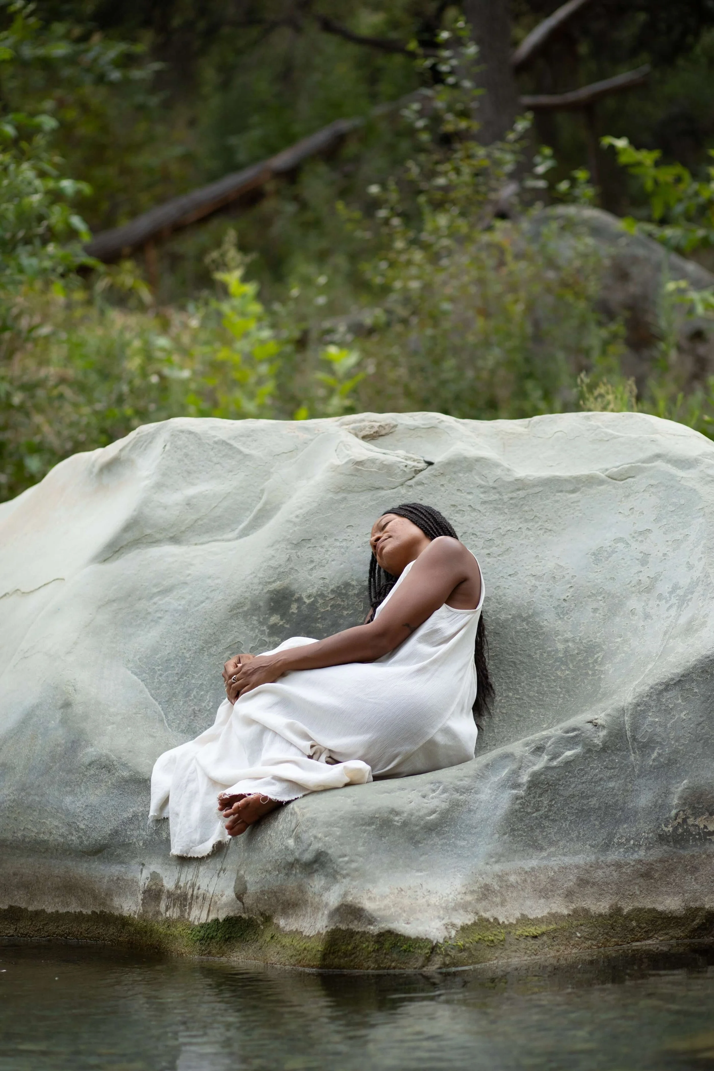 A woman in a white dress sitting on a large rock by a body of water in a forest, with her head tilted back and eyes closed.