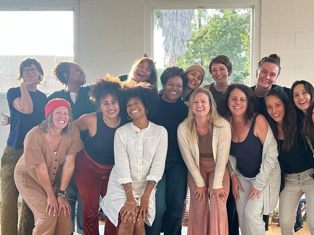 Group of women and non-binary folks smiling and posing together indoors near large windows.