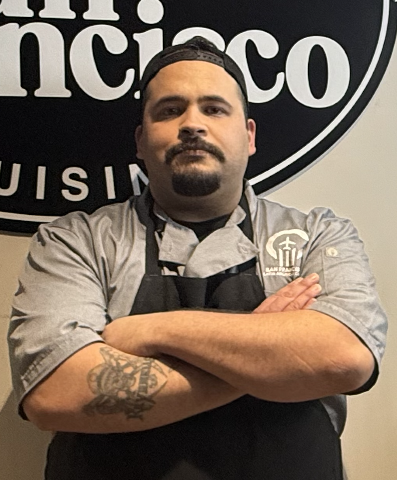 A chef with crossed arms wearing a gray chef coat and a black apron, standing in front of a black and white sign that reads 'Tamicoco.'