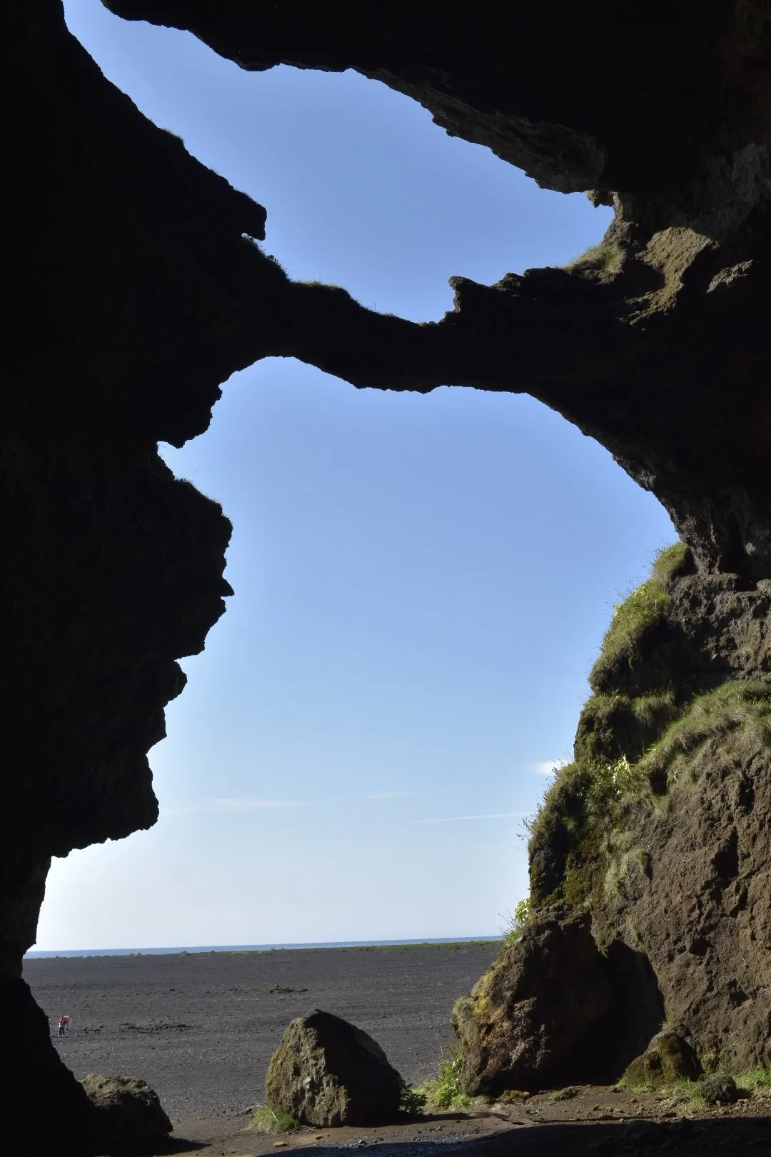 View through a volcanic rock formation looking out onto a flat black sand landscape with the ocean in the distance, under a clear blue sky.