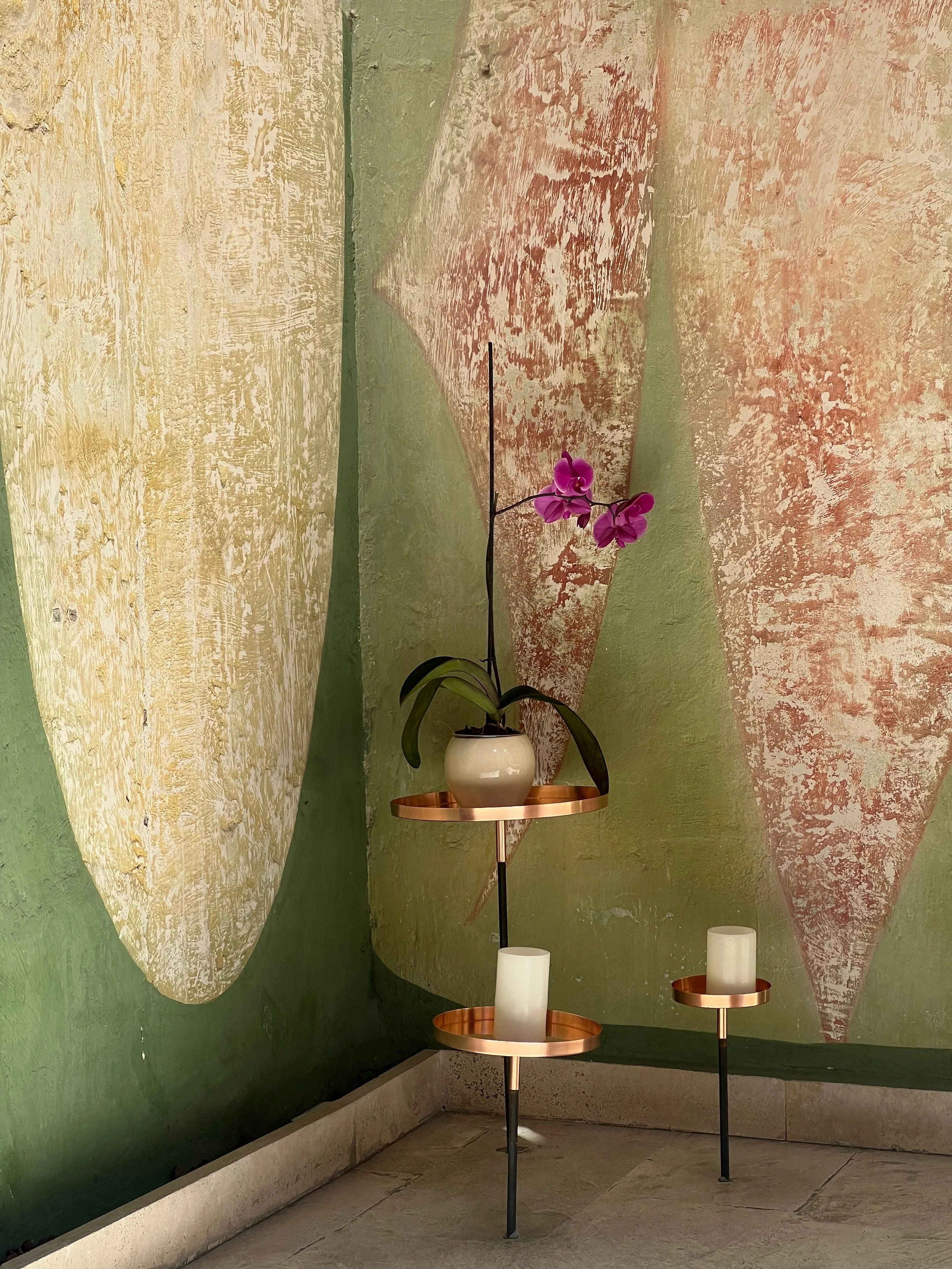 A decorative plant stand with an orchid in a white pot on the top tier and two white candles on the lower tiers, against a textured wall with abstract painted shapes.