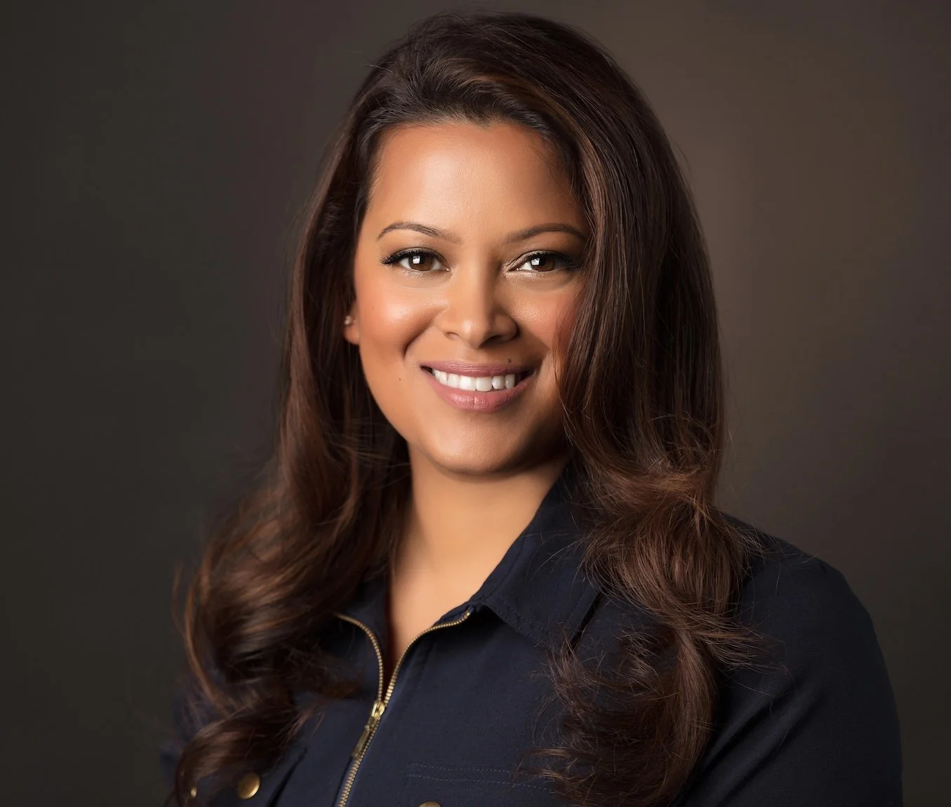 Close-up portrait of a smiling woman with long brown hair, wearing a dark jacket with a zipper, against a dark background.