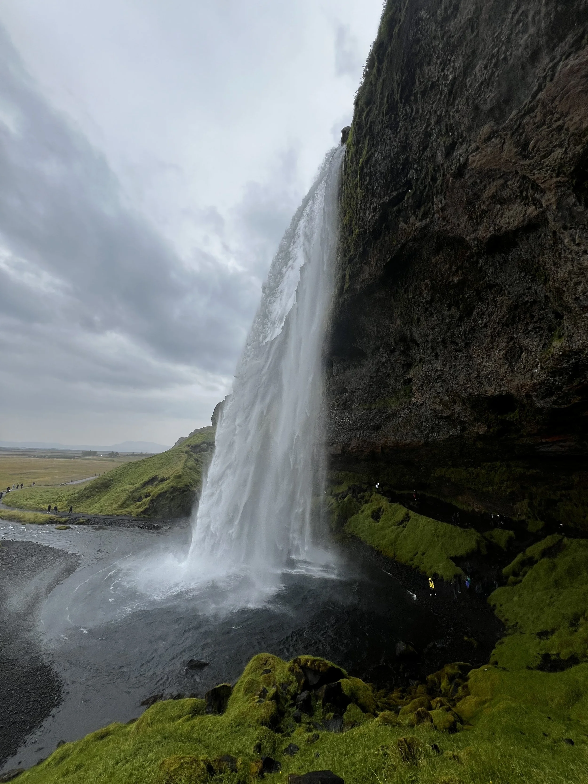 A tall waterfall flowing over a dark cliff into a pool below, with green moss and grass surrounding the base and cloudy sky overhead.