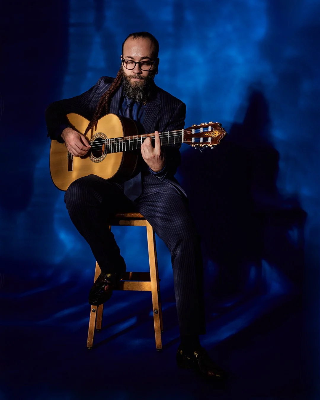 A man in a pinstripe suit with glasses and a beard, sitting on a wooden stool, playing an acoustic guitar against a blue background.
