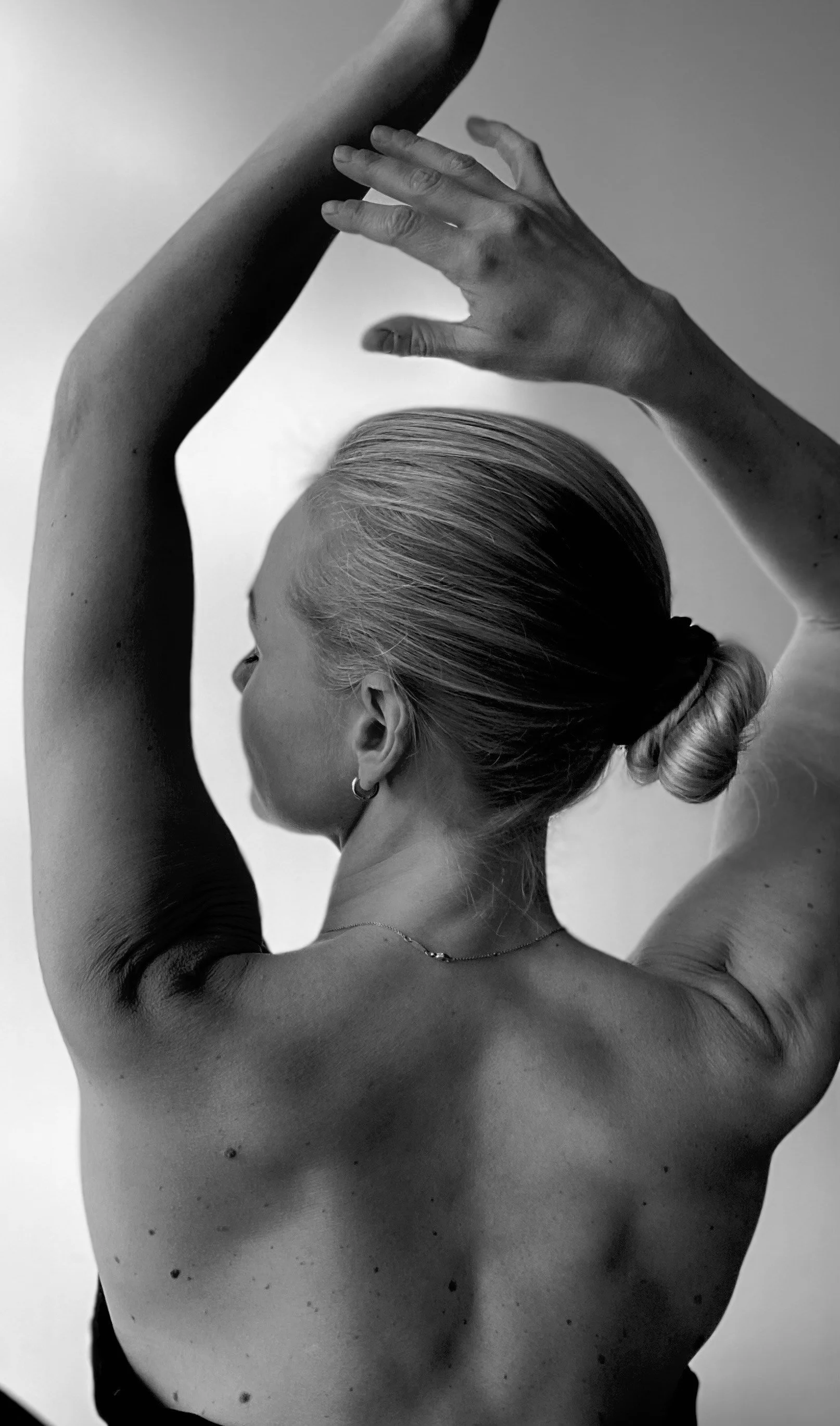 Black and white image of a woman with her hair in a bun, viewed from the side, raising her arms above her head in a dance or yoga pose.