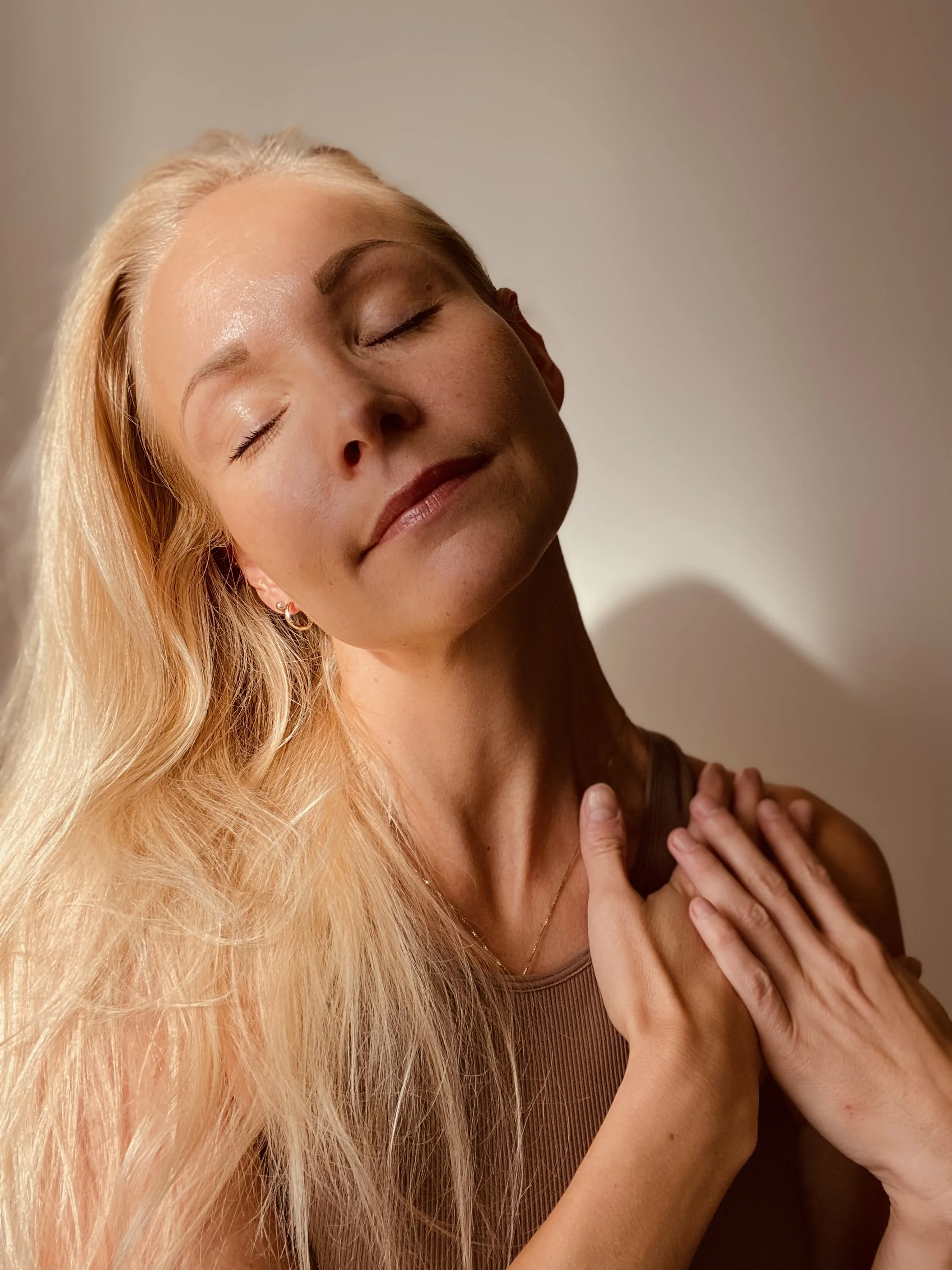 A woman with long blonde hair and closed eyes, smiling gently, holding her hands over her chest.