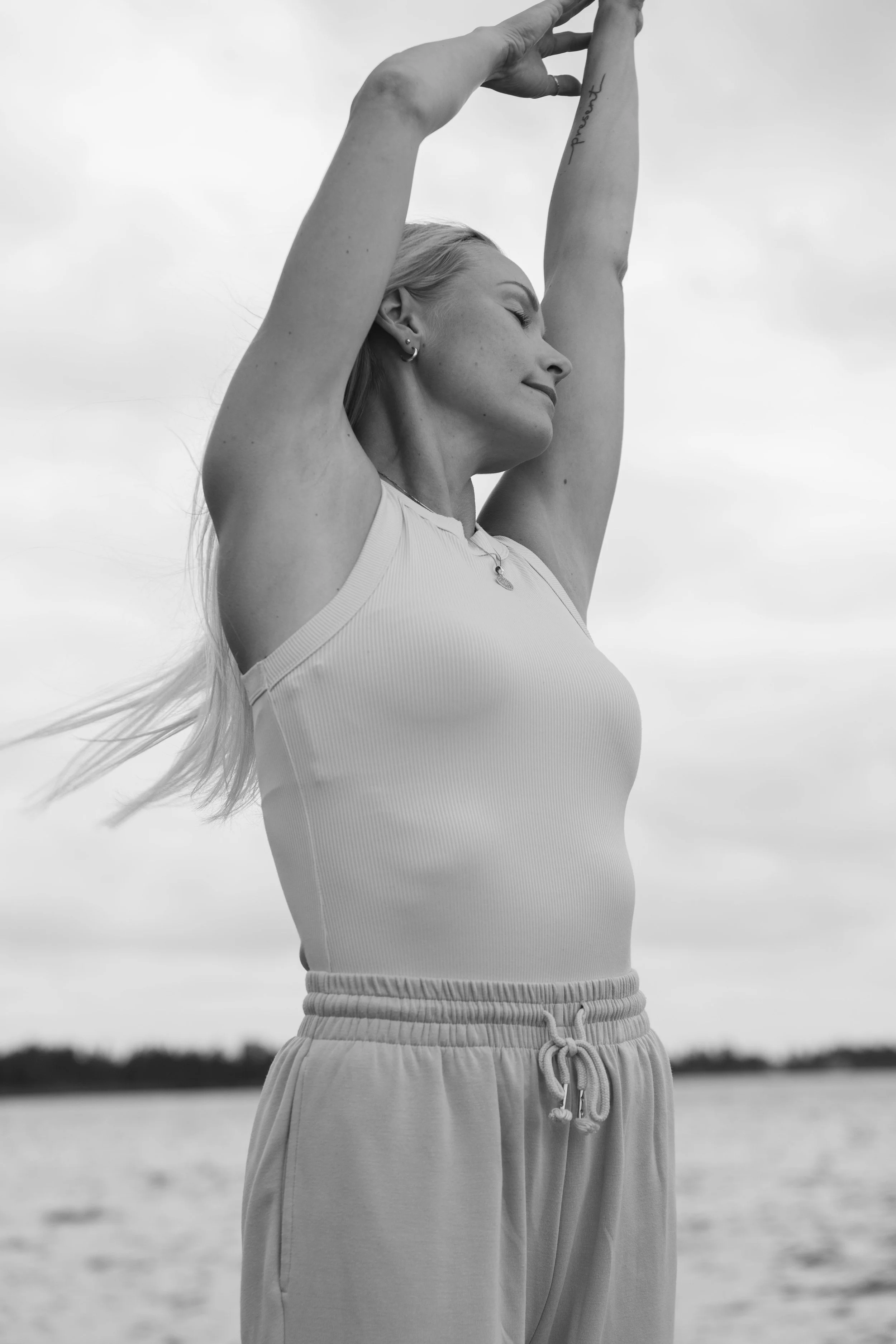A woman with long hair in a sleeveless top and sweatpants, standing outdoors with her eyes closed and arms raised above her head, relaxing by a body of water.