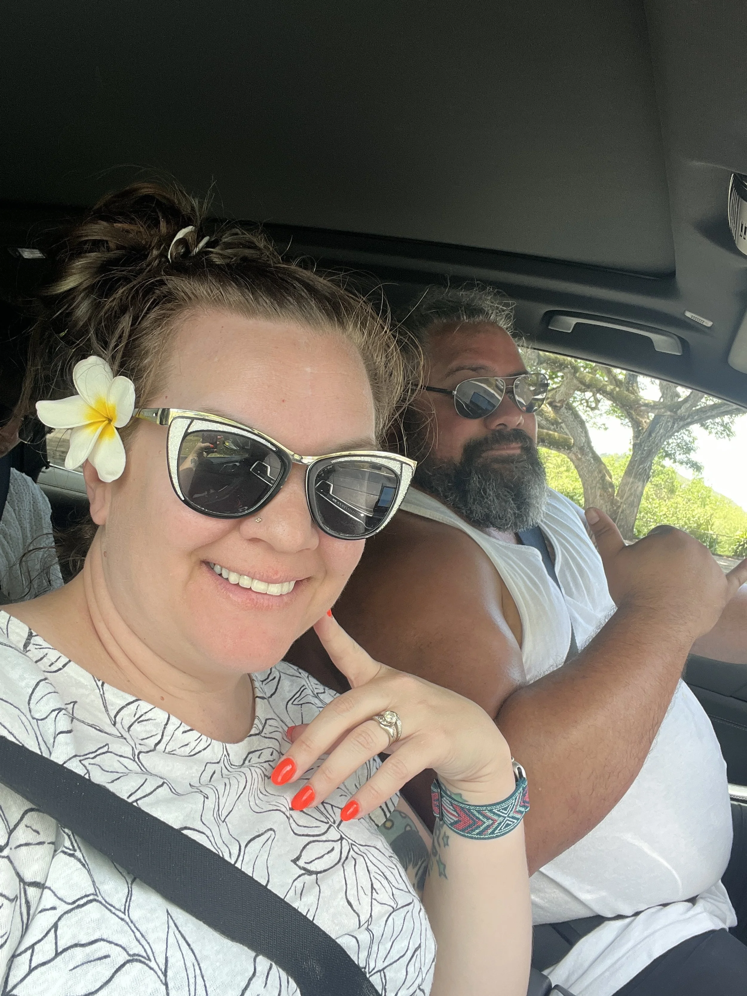 A woman and a man sitting in a car, both wearing sunglasses. The woman is smiling, with a white flower in her hair and red nails, pointing to her face. The man is giving a shaka hand sign and has a beard and gray hair.