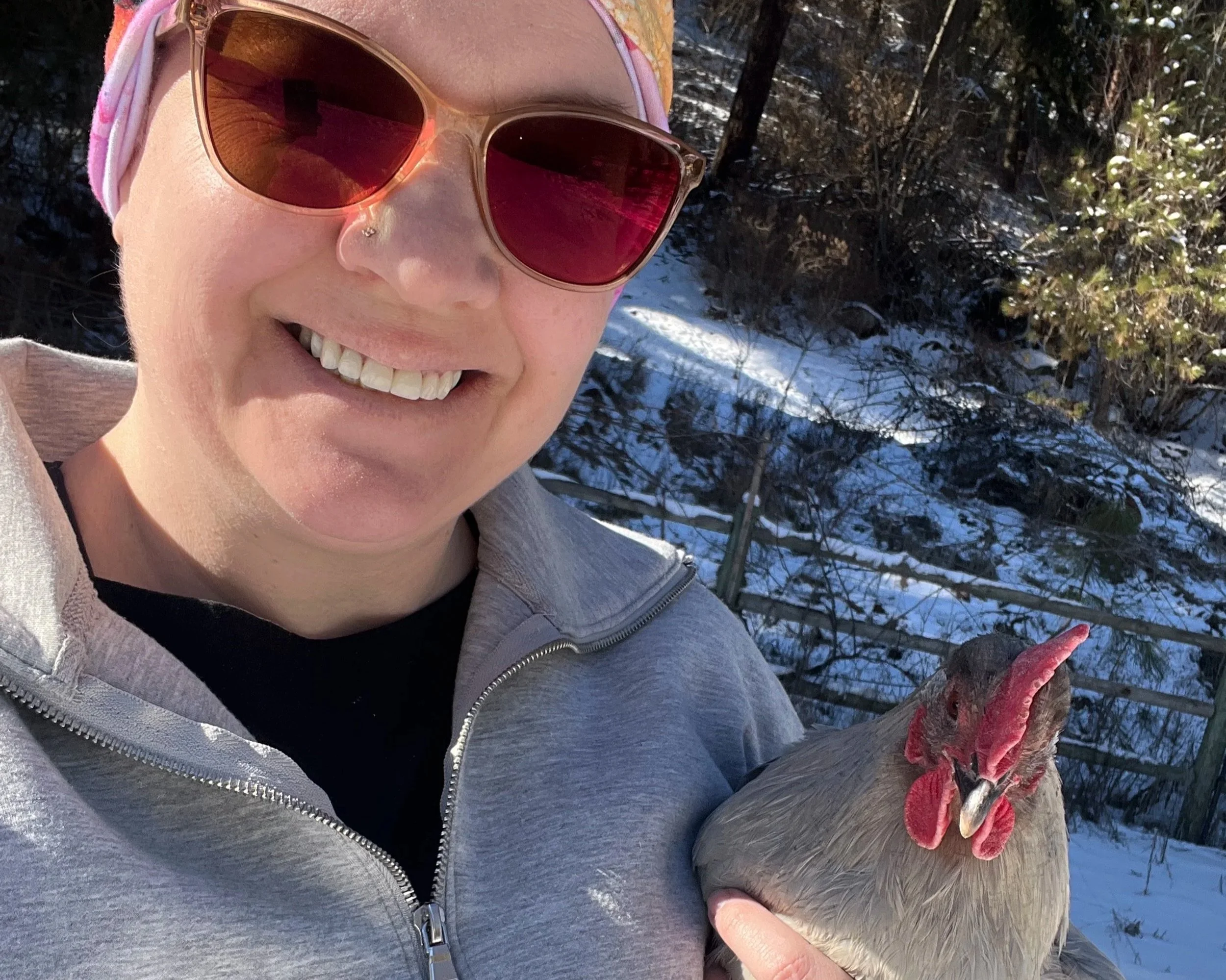 A woman smiling while holding a chicken outdoors in a snowy area, wearing sunglasses, a pink headband, and a gray zip-up jacket.