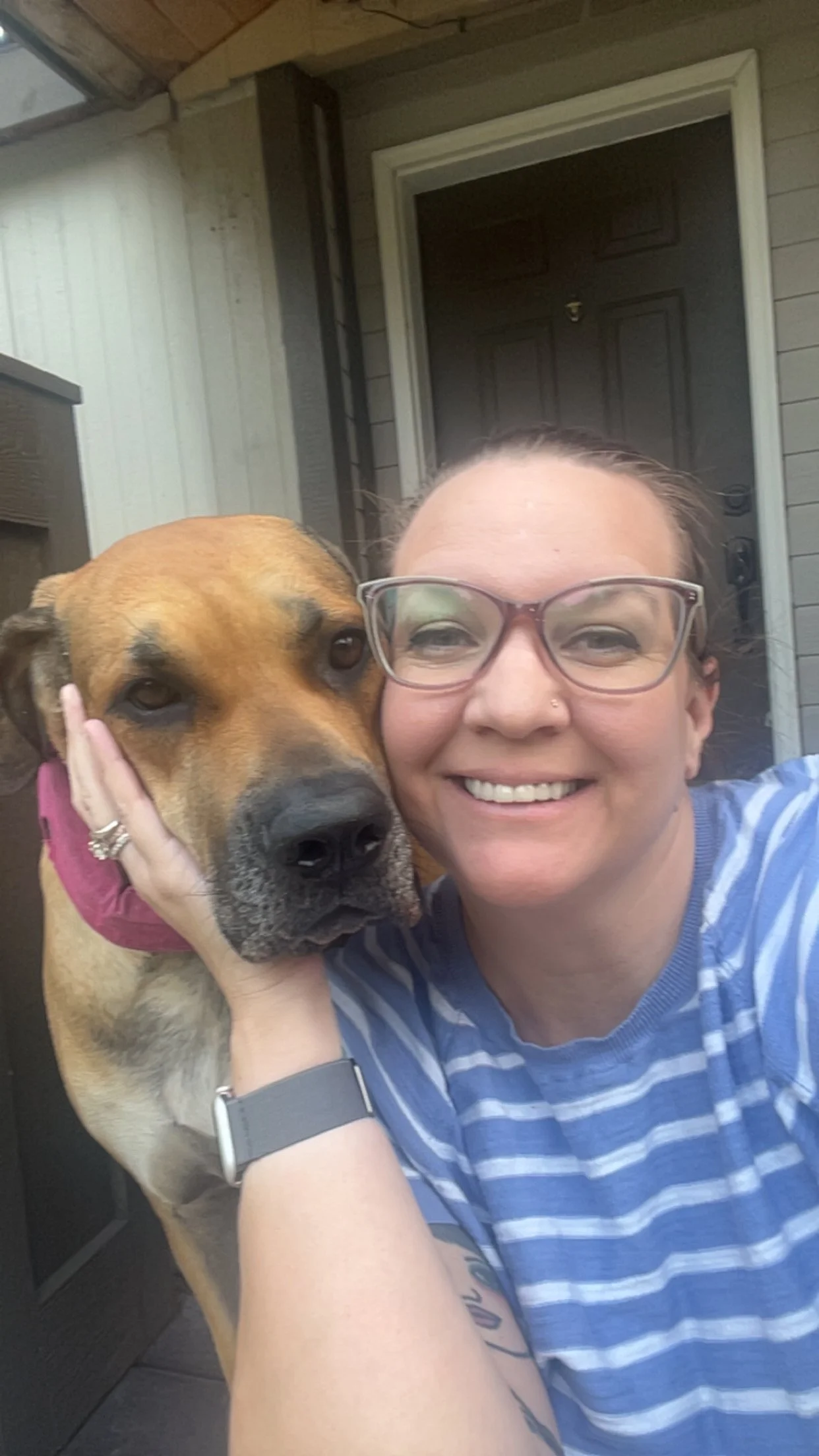 A woman with glasses smiling and hugging a large brown dog with a pink collar outside a house, in front of a door.