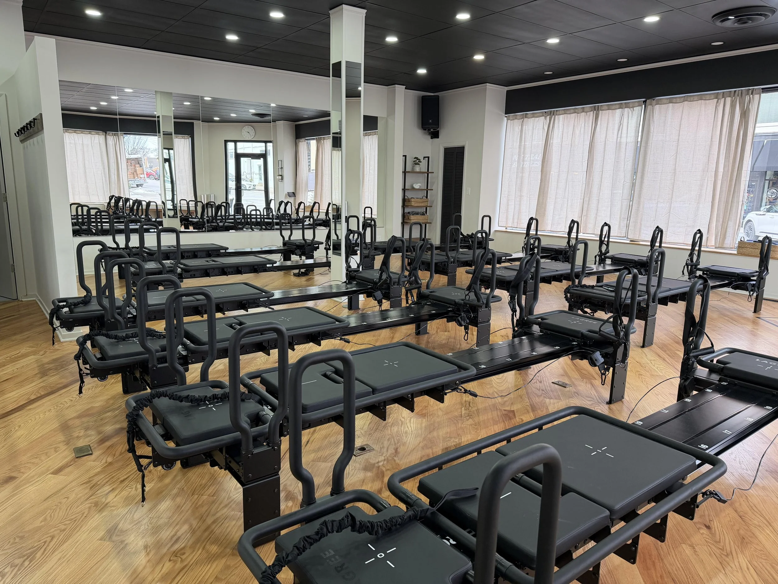 Pilates reformer machines arranged in a fitness studio with wooden floors, large mirrors, windows with curtains, and ceiling lights.