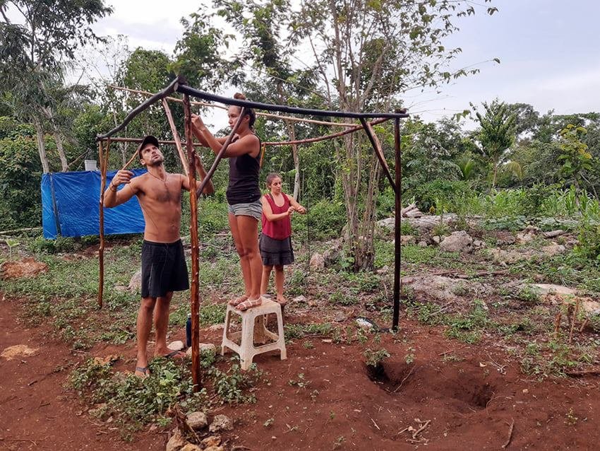 Three people working together to set up a makeshift canopy frame outdoors, with one person standing on a stool adjusting the top pole, in a rural area with trees and dirt ground.