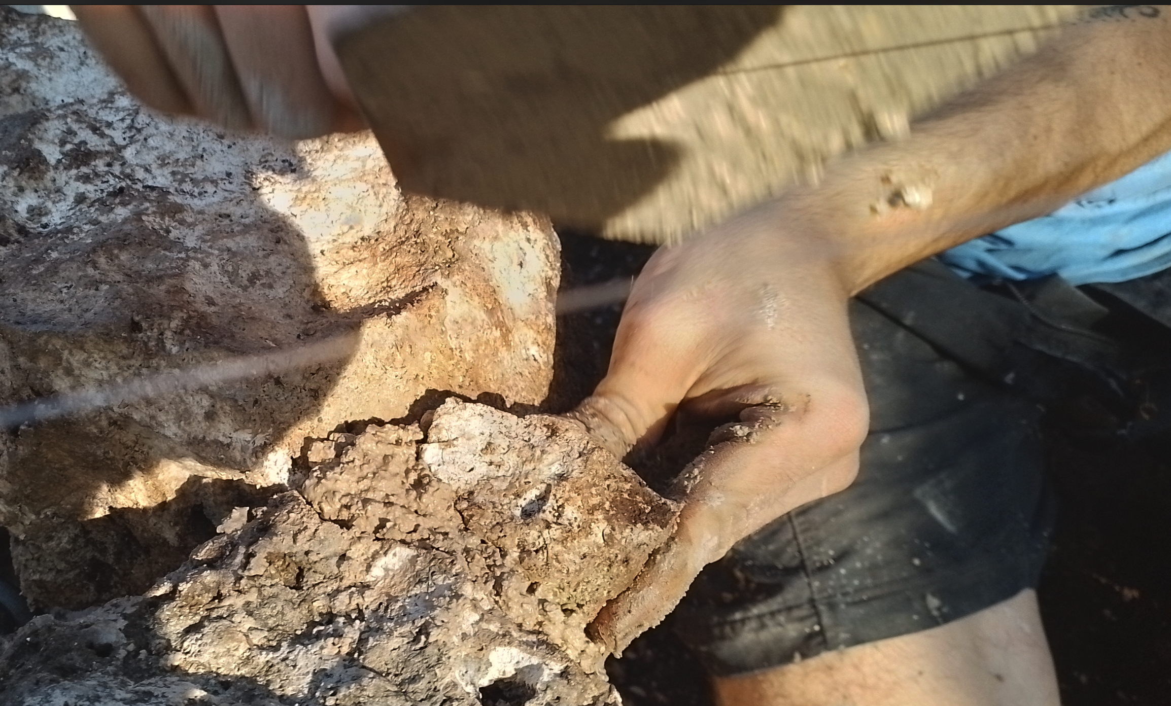Close-up of a person's hand working with a rough, light-colored stone or rock. The person is wearing a blue garment and appears to be involved in an activity like construction or excavation, with dirt visible on the skin and clothing.